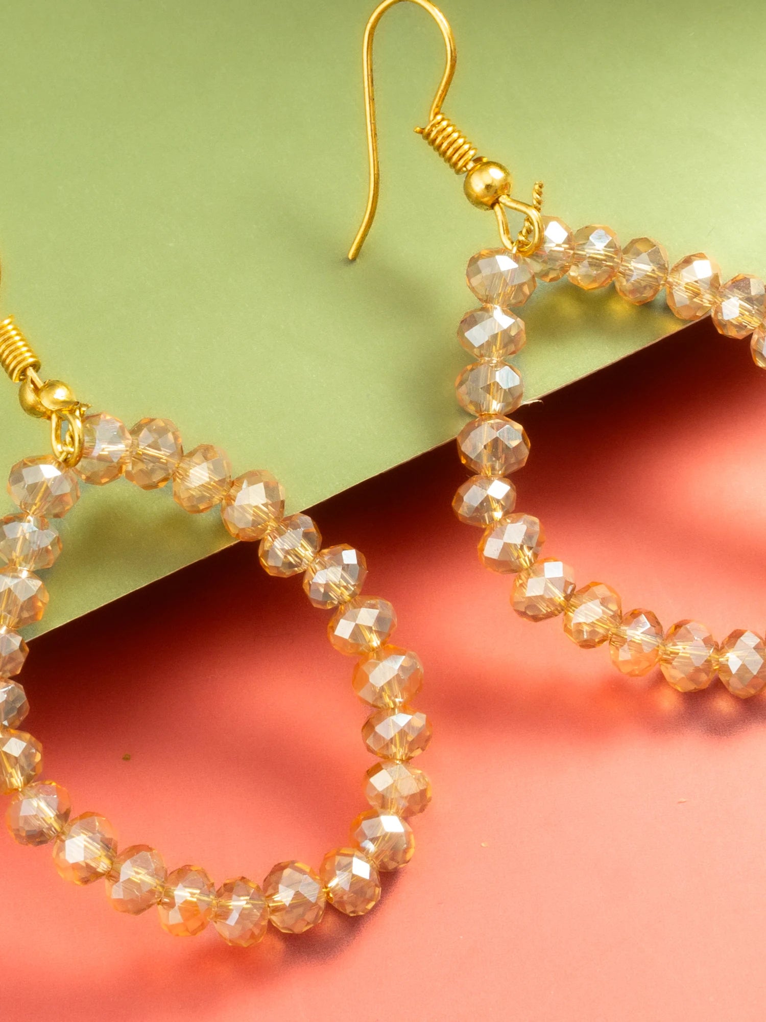 Gold-tone beaded hoop earrings with faceted clear beads, shown on a split green and coral background.