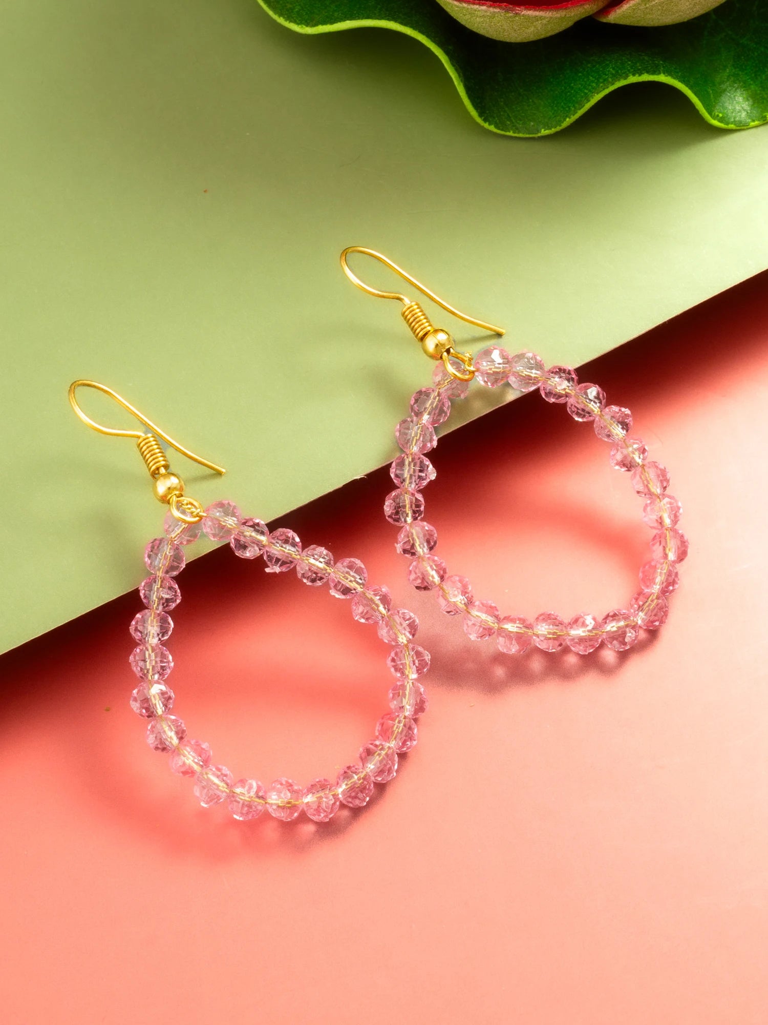 Pink faceted bead hoop earrings with gold fishhook ear wires, displayed on a split green and coral background with a green leaf at the top.