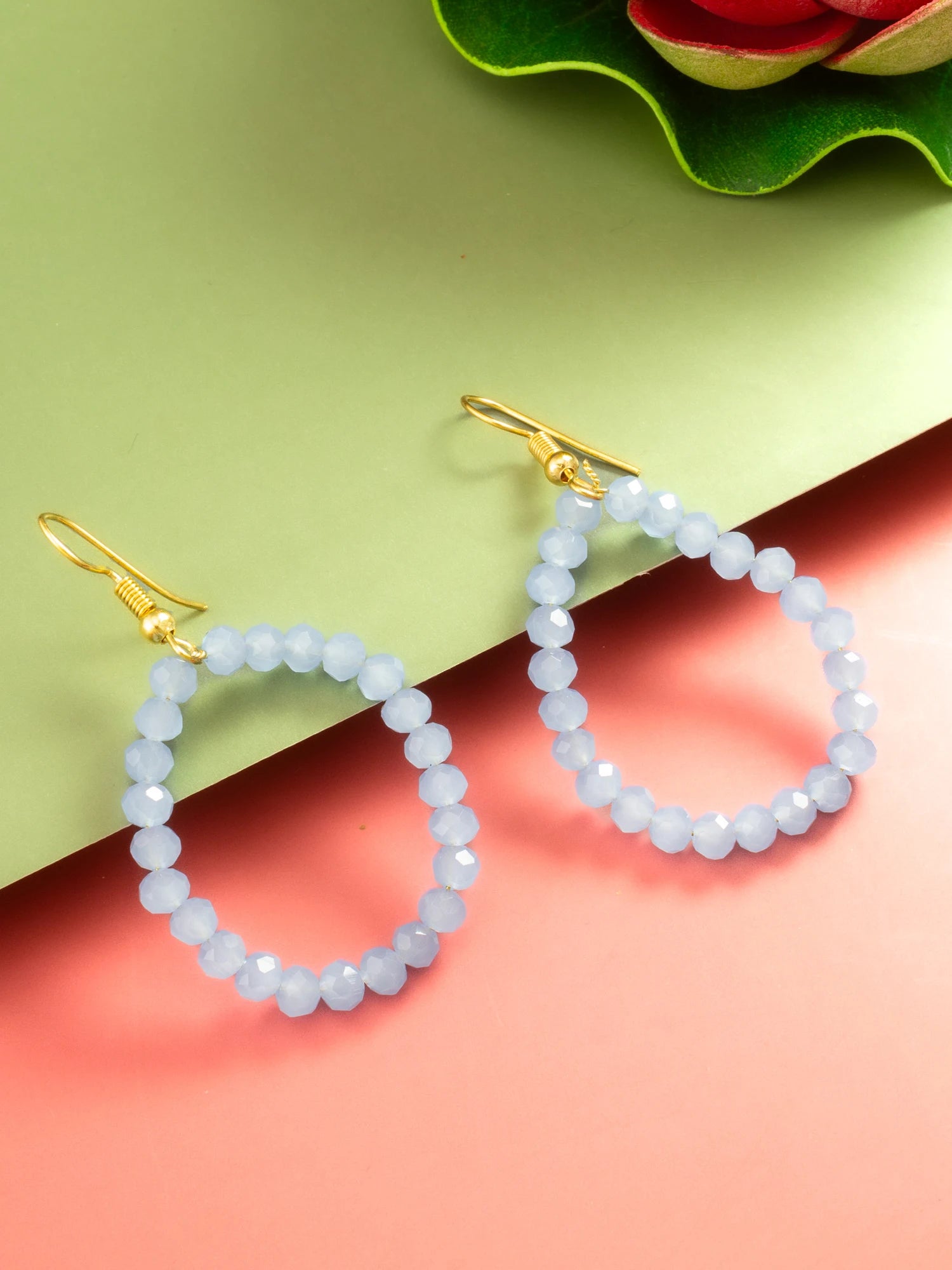 A pair of gold-tone drop earrings with faceted pale blue beads, displayed on a split green and pink background with a decorative green leaf and red flower in the corner.