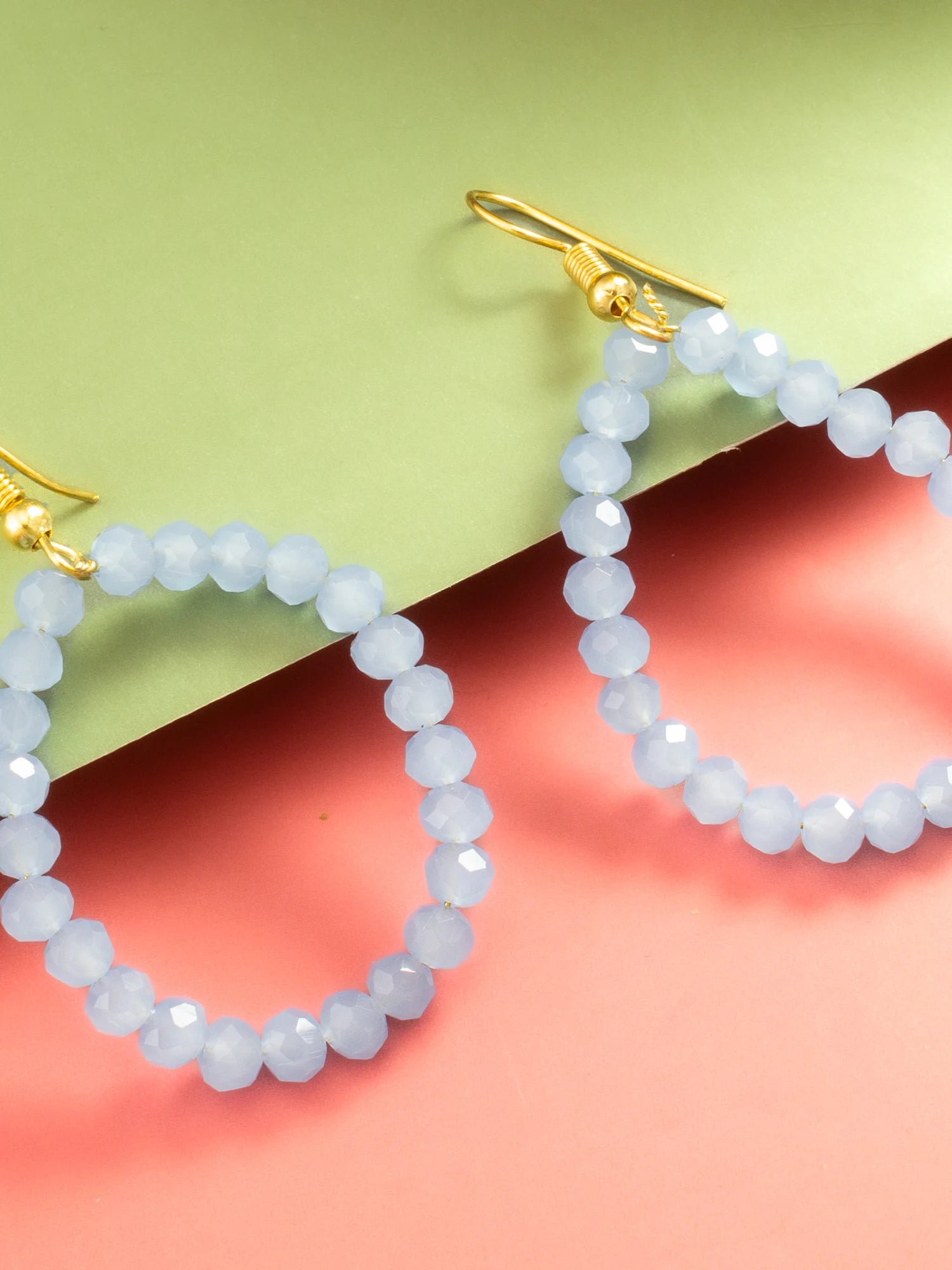 Light blue faceted bead hoop earrings with gold fishhook ear wires, displayed on a split pastel green and coral pink background.