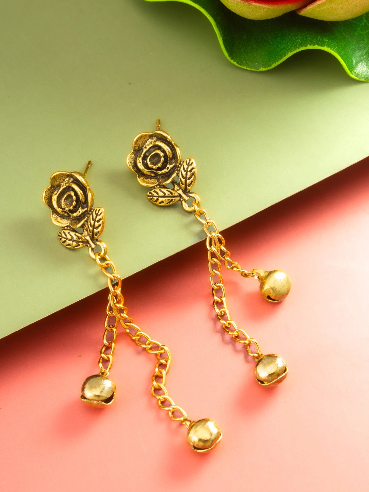 A pair of gold-toned rose-shaped dangle earrings with leaf details and small bell-shaped charms, displayed on a split green and pink background.