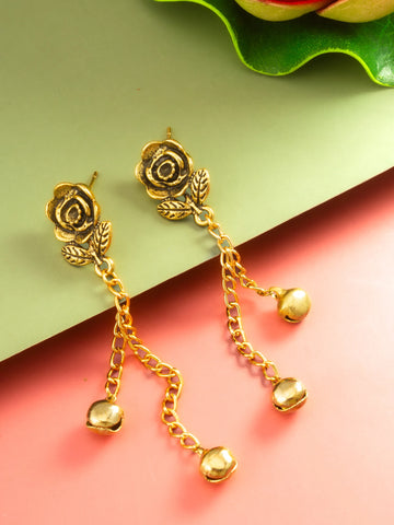 A pair of gold-toned rose-shaped dangle earrings with leaf details and small bell-shaped charms, displayed on a split green and pink background.