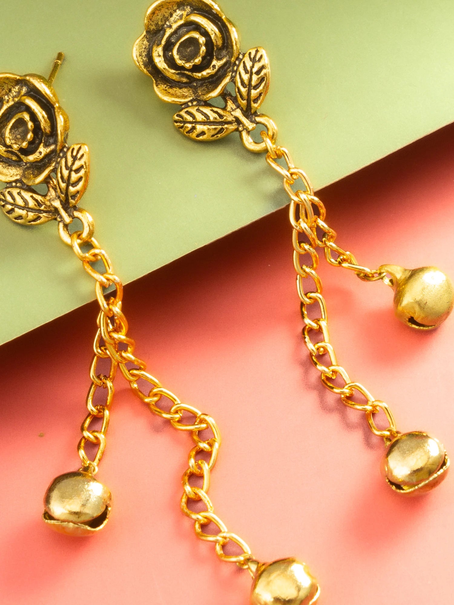 Gold rose dangle earrings with leaf details and small bell charms, shown on a split green and pink background.