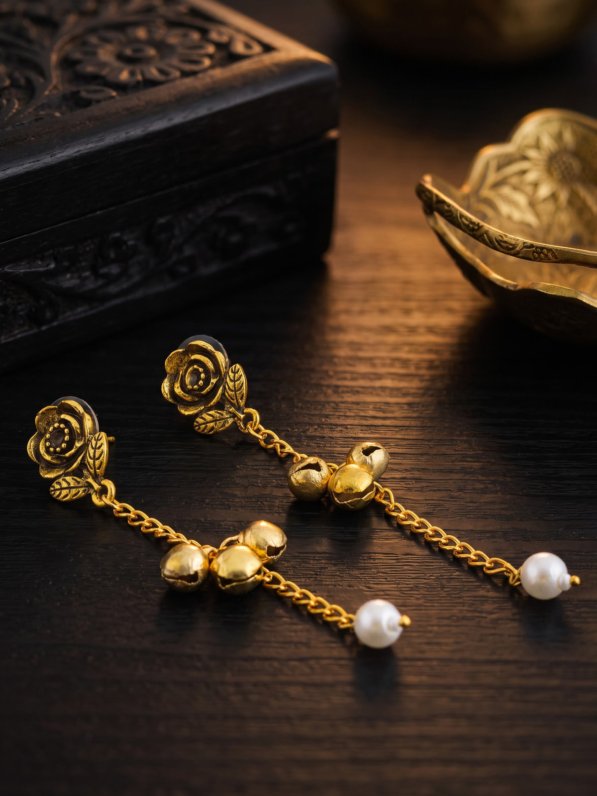 A pair of gold-toned dangle earrings with rose-shaped studs, small bell charms, chain links, and white pearl drops, displayed on a dark wooden surface with decorative objects in the background.