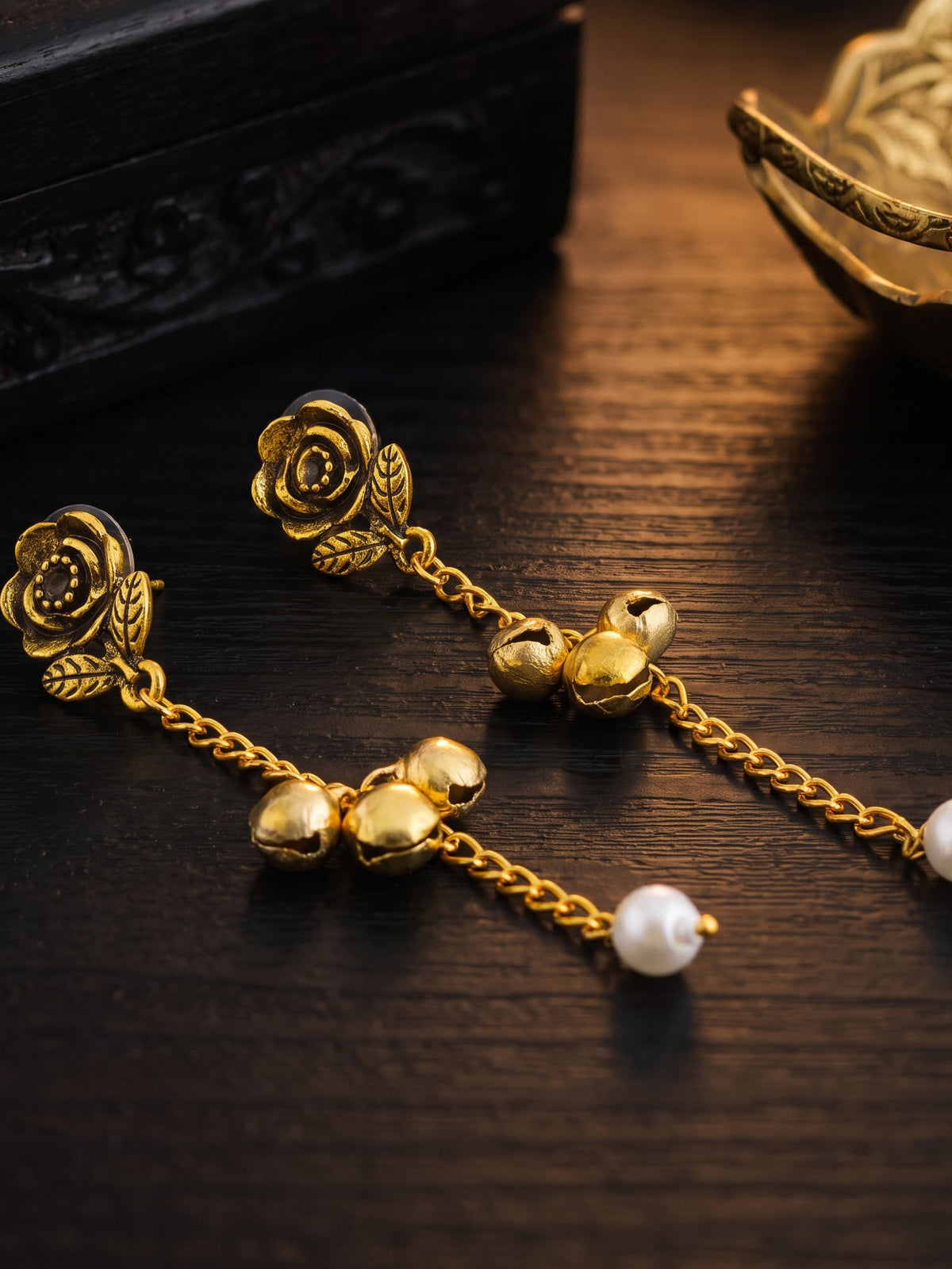 A pair of gold-toned dangle earrings with rose-shaped studs, small bell charms, chain links, and white pearl beads, displayed on a dark wooden surface.
