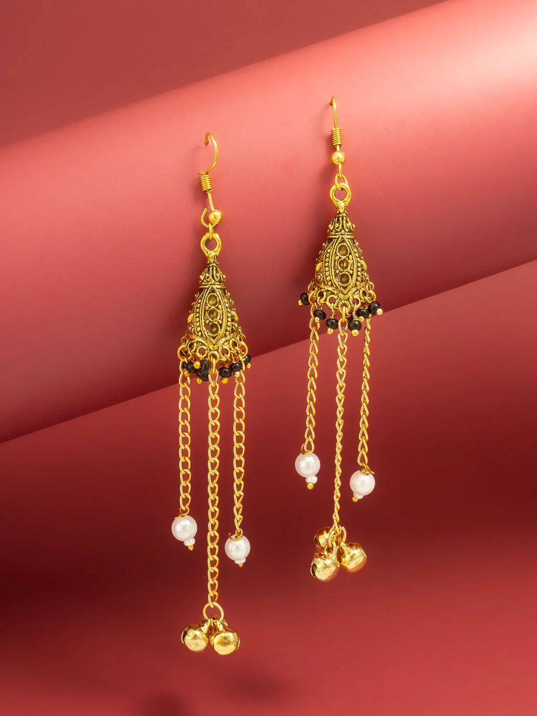A pair of gold-toned Indian-style dangle earrings with intricate filigree work, small black bead accents, and dangling white pearl-like beads on a pink background.