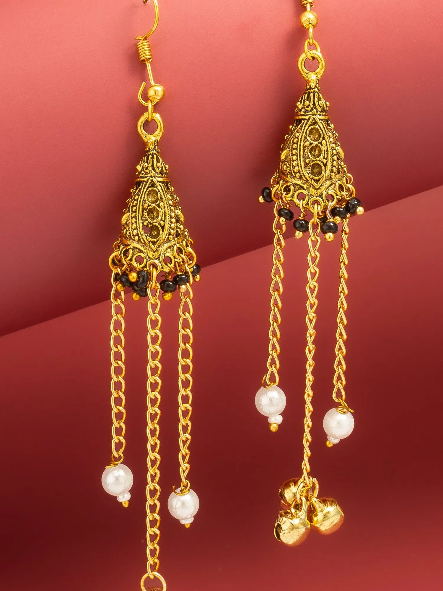 A pair of gold-toned traditional Indian jhumka earrings with intricate filigree work, small black bead accents, and dangling white pearl beads, set against a solid dark red background.