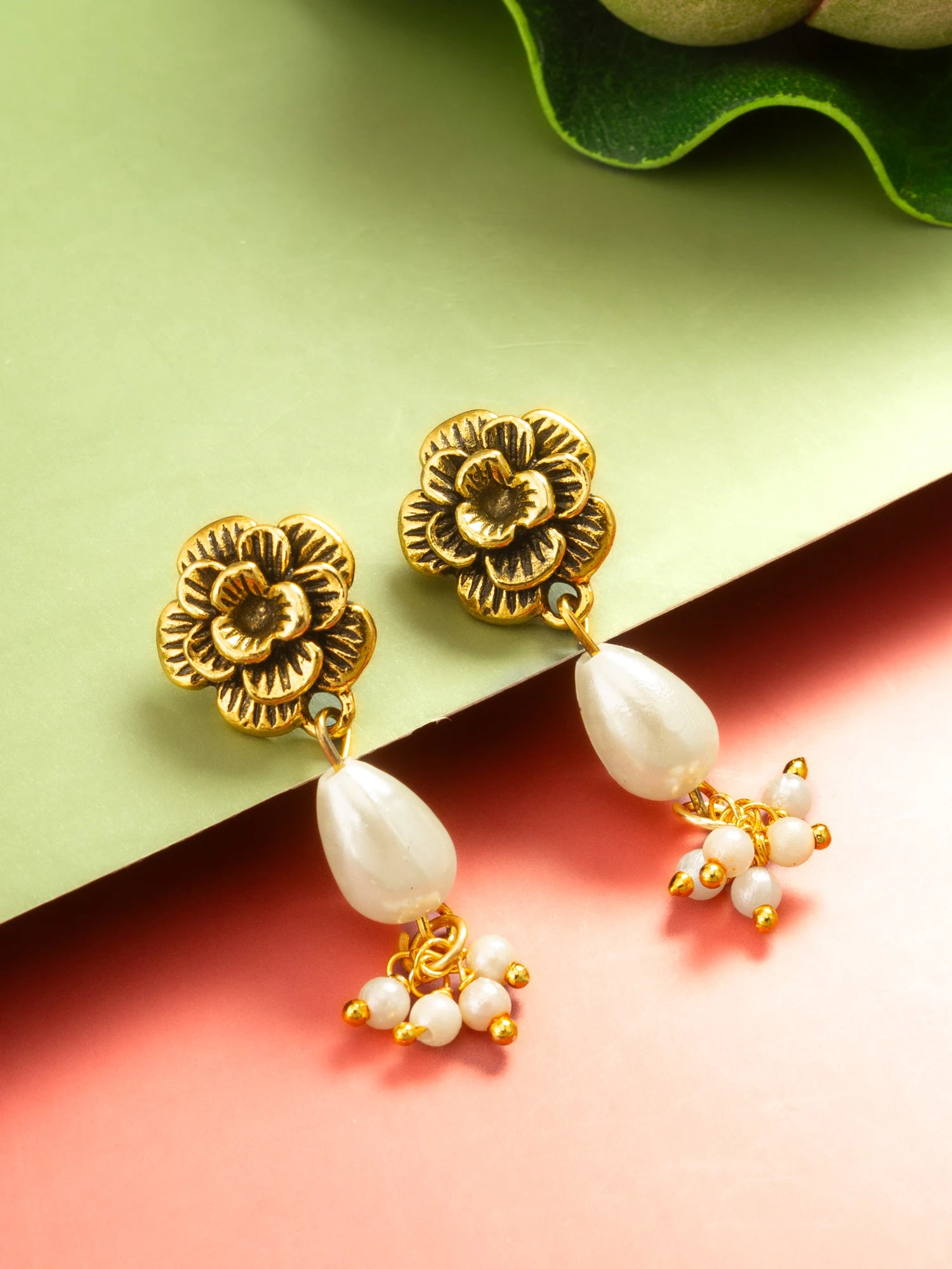 A pair of gold-toned dangle earrings with embossed floral tops, white teardrop pearls, and small pearl clusters, styled on a split green and pink background with a leaf in the corner.