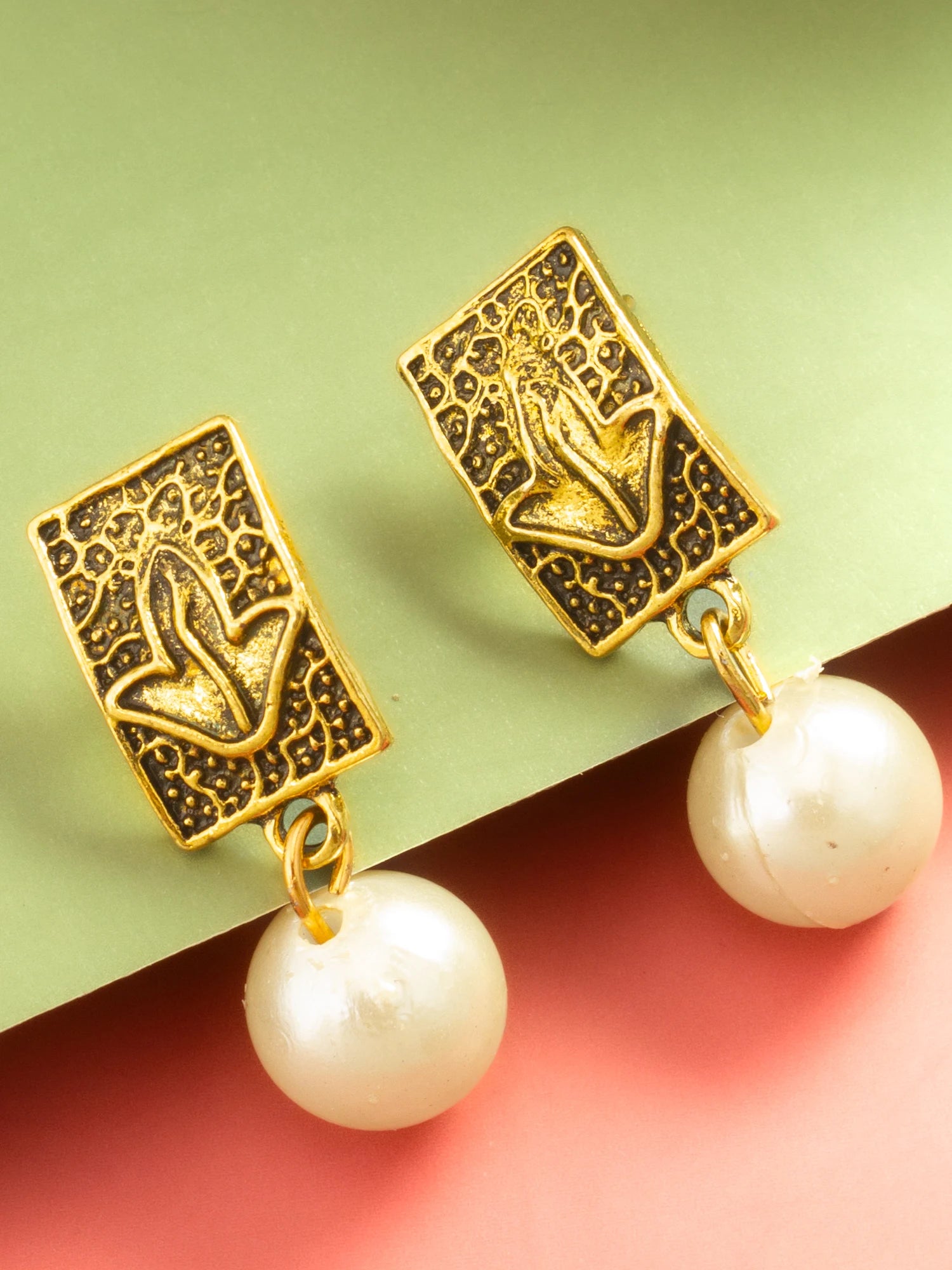 A pair of gold-toned rectangular drop earrings with textured filigree detailing and dangling white pearl beads, displayed on a split green and pink background.