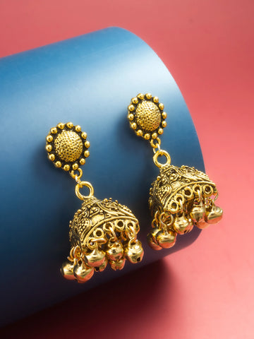 A pair of gold-toned Indian jhumka earrings displayed on a blue cylindrical prop against a pink background.