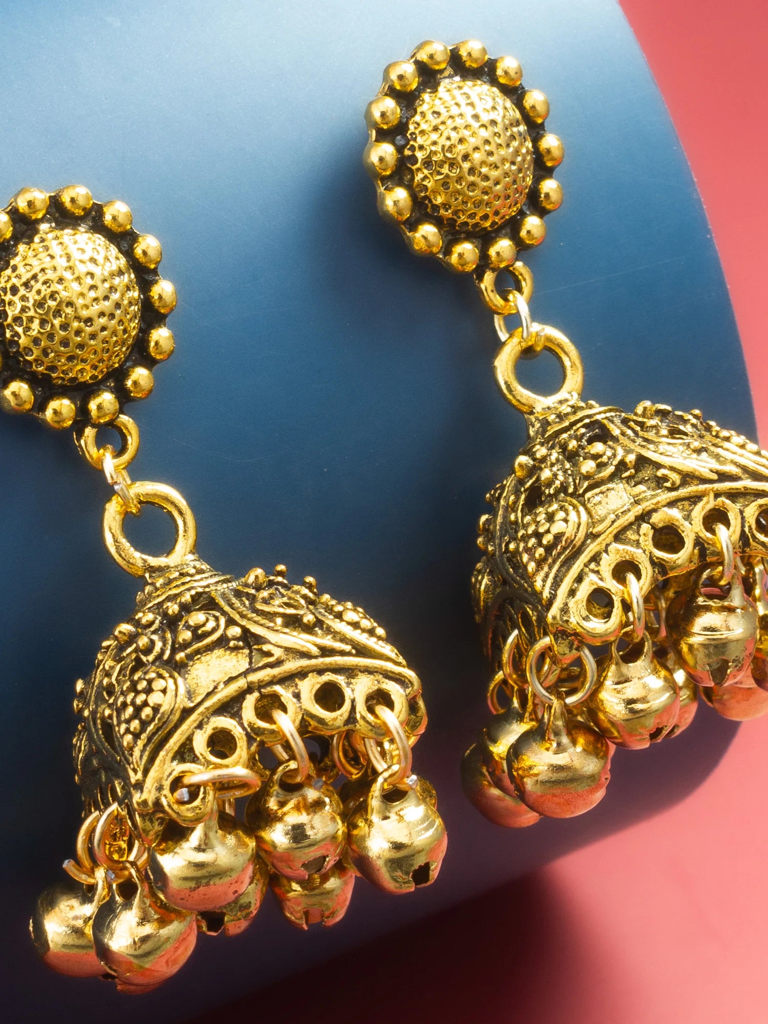 A pair of ornate gold-toned jhumka earrings with detailed filigree work and dangling bells, displayed on a blue and pink background.