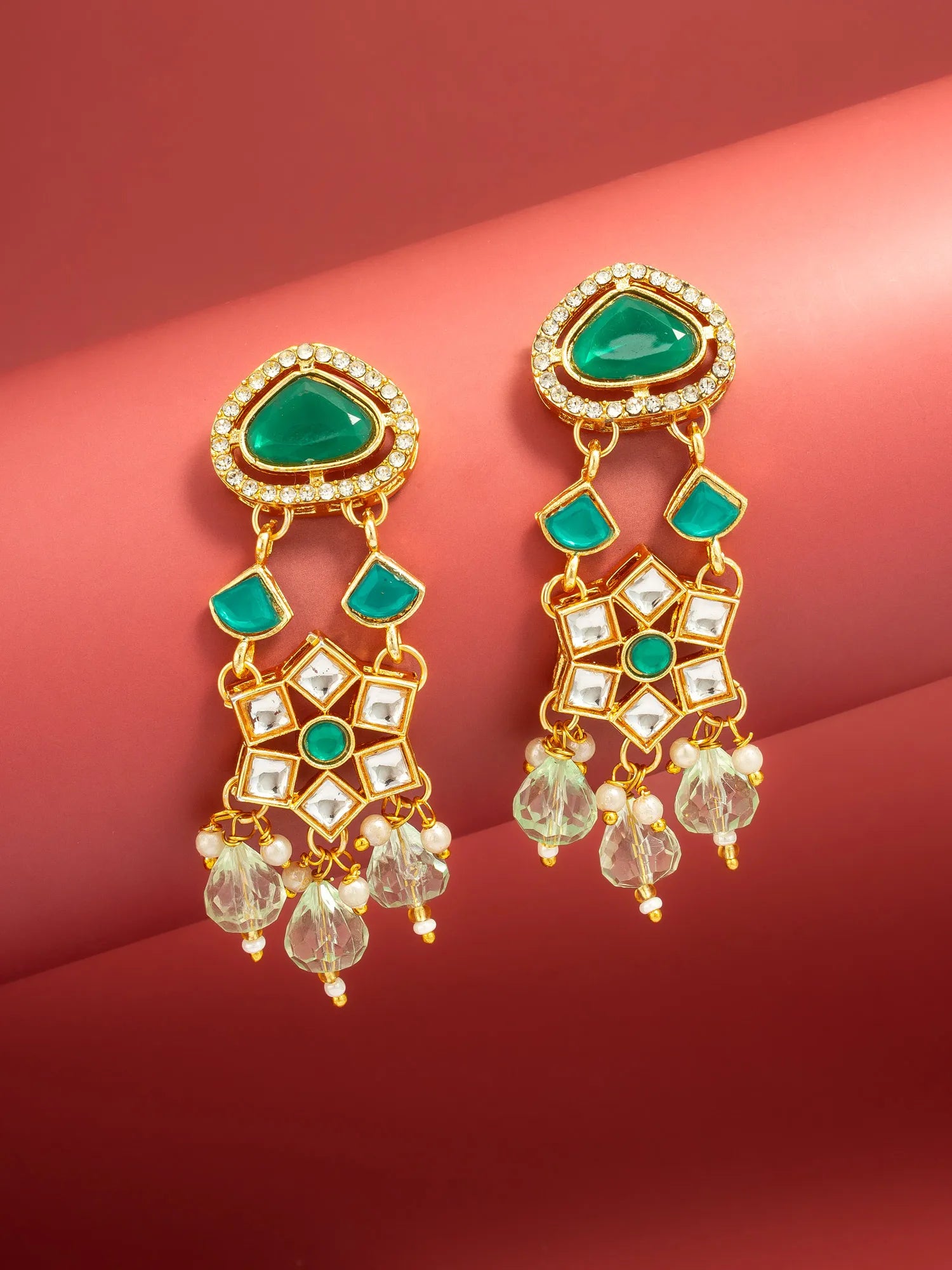 A pair of gold-plated dangle earrings with green stone accents and clear crystal drops, displayed on a red background.