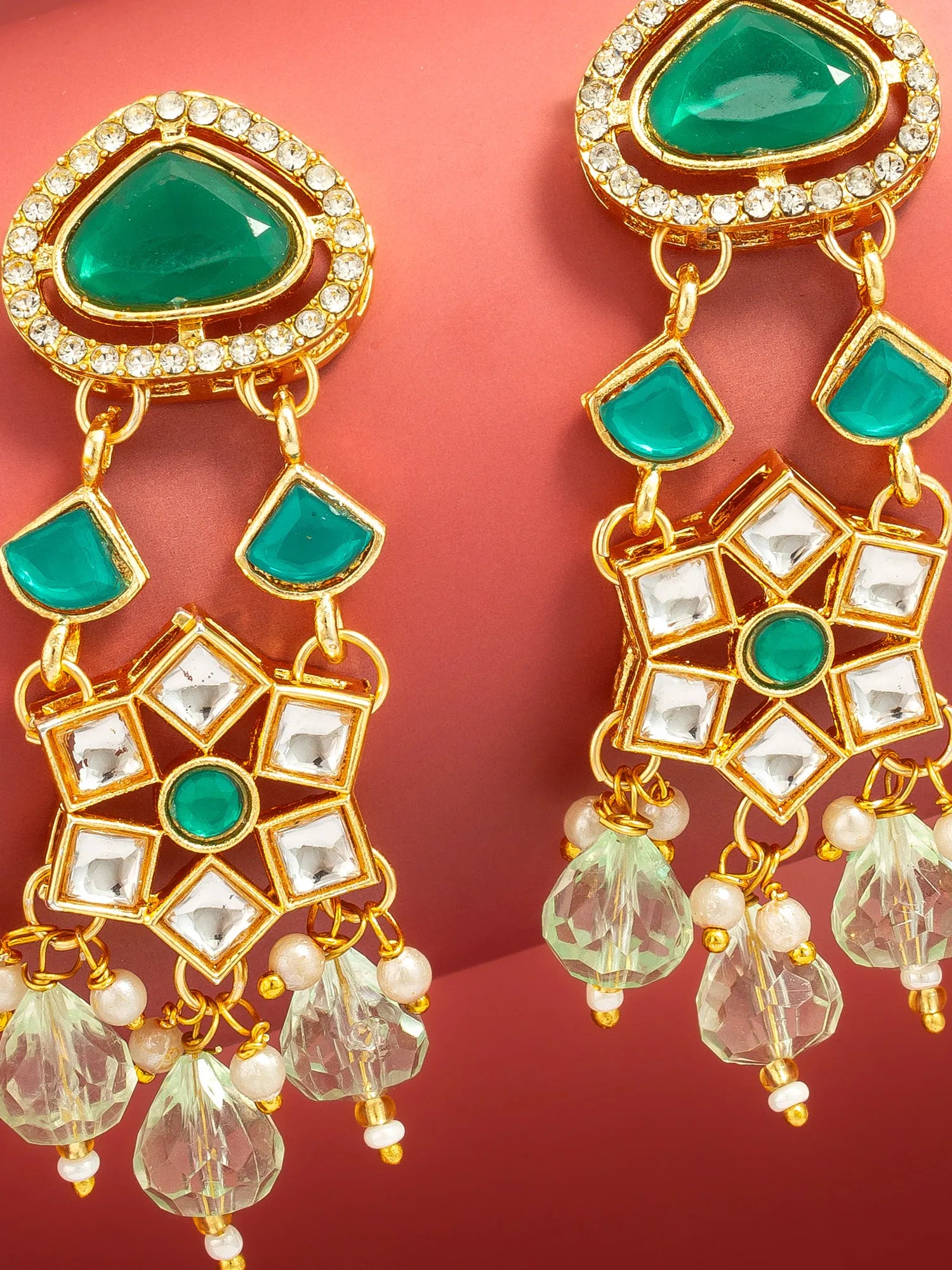 A pair of gold-plated Indian kani earrings with green stones and clear crystal accents, displayed against a solid red background.