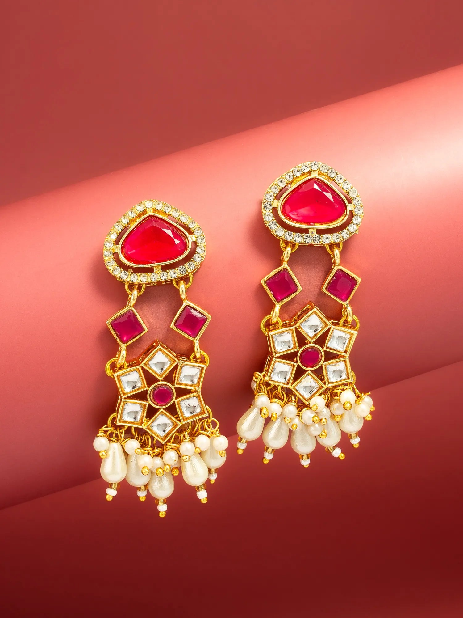 A pair of gold-plated Indian jhumka earrings featuring red inlay stones, clear crystal accents, and white pearl drops, displayed on a curved red background.
