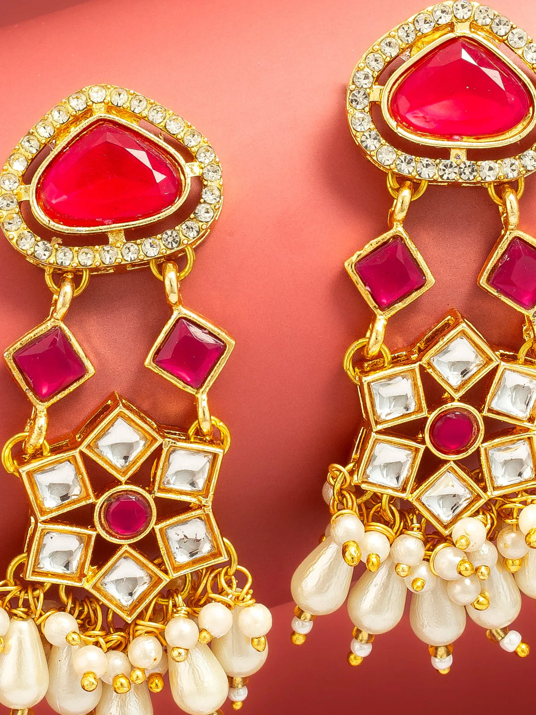 A pair of gold-plated Indian jhumka earrings featuring red inlay stones, clear crystal accents, and white pearl drops, displayed on a curved red background.