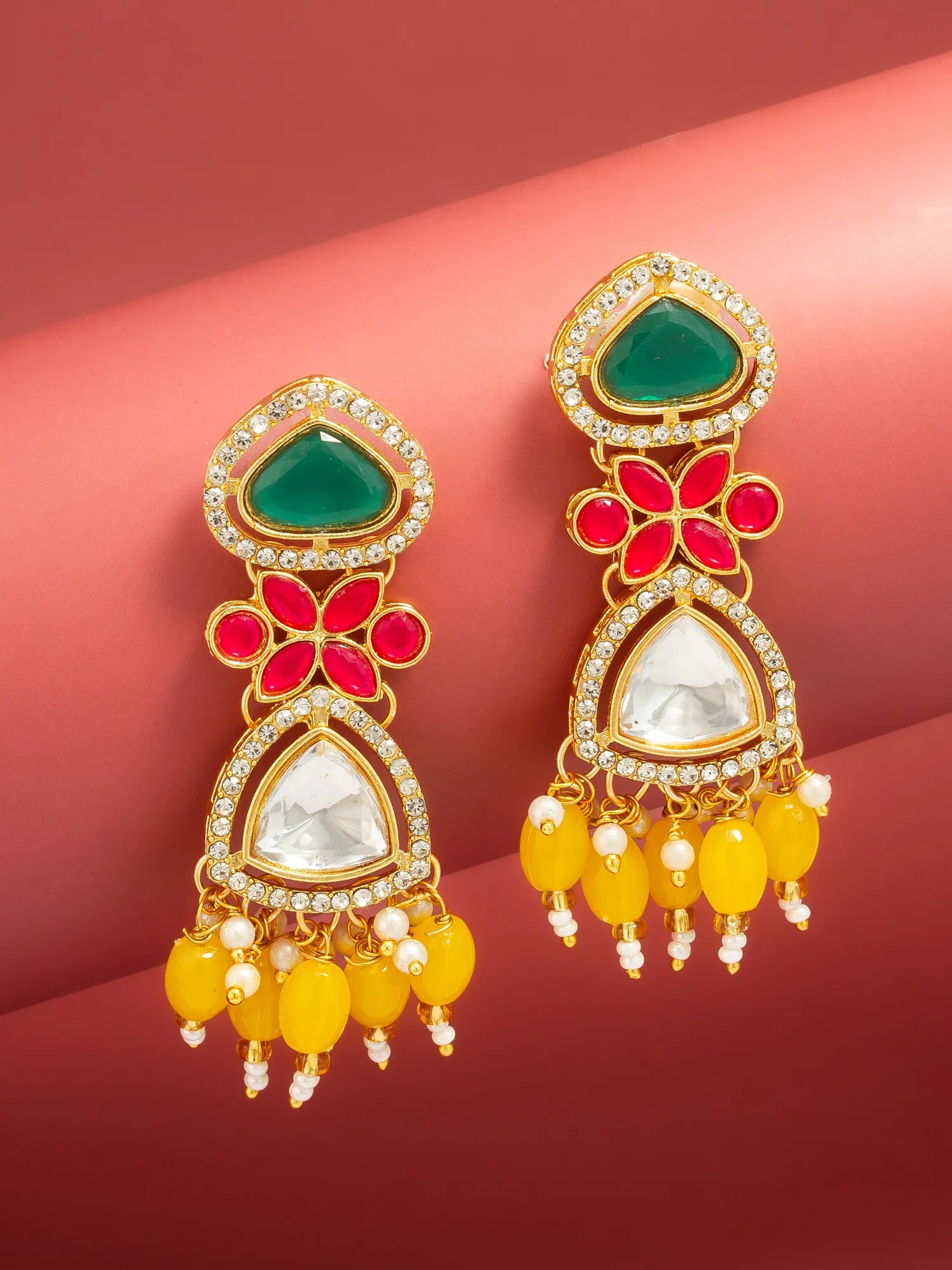 A pair of gold-plated Indian jhumka earrings with green, red, and white stones, accented with yellow beads and small white pearls, set against a pink background.
