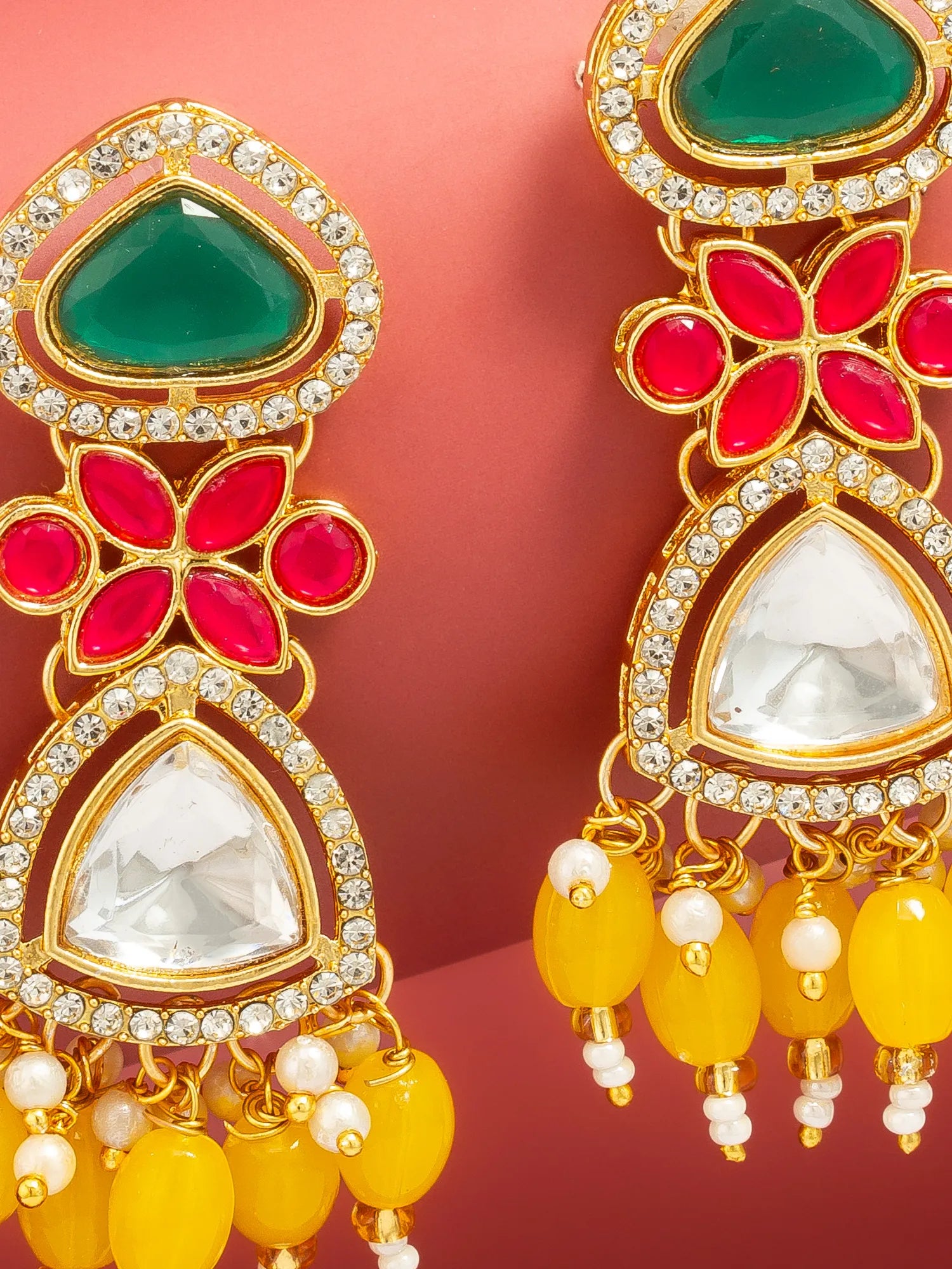 Gold-plated statement earrings with green, red, and white stones, accented with clear crystals and yellow bead tassels, shown against a pink background.