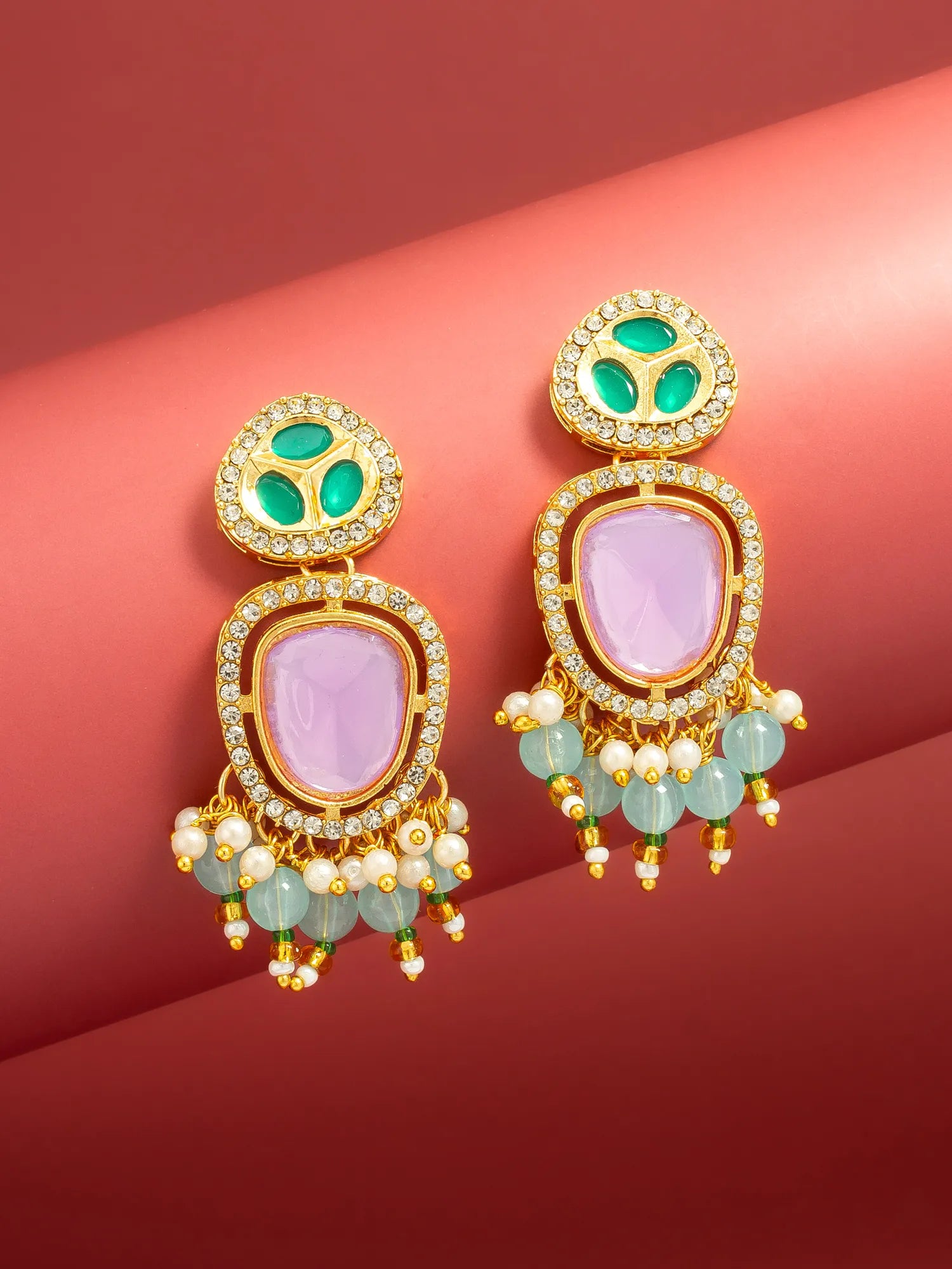 A pair of ornate gold-plated drop earrings with light purple oval stones, green enamel details, and dangling light green beads and white faux pearls, displayed on a curved red background.