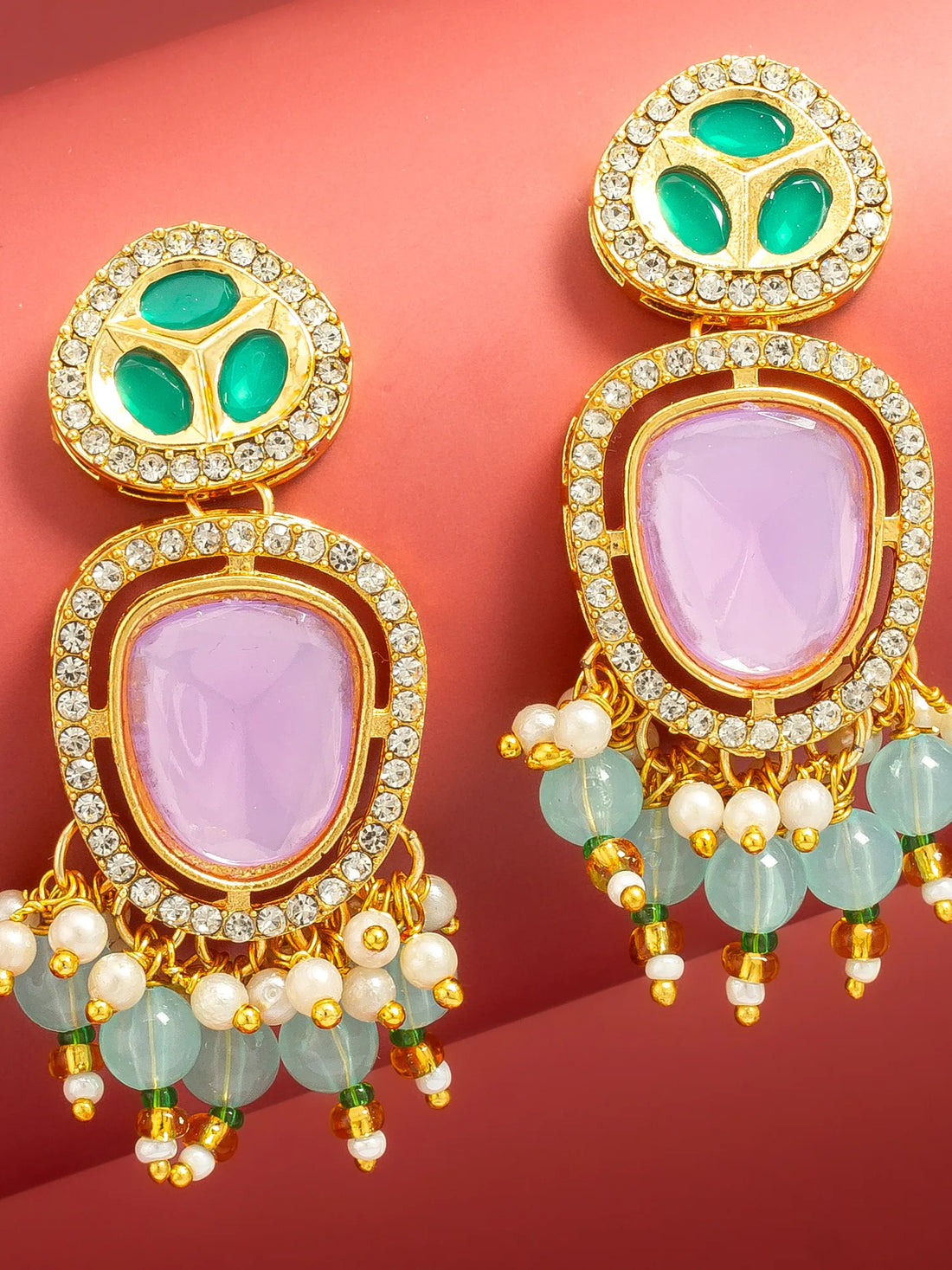 A pair of ornate gold-plated drop earrings with light purple oval stones, green enamel details, and dangling light green beads and white faux pearls, displayed on a curved red background.