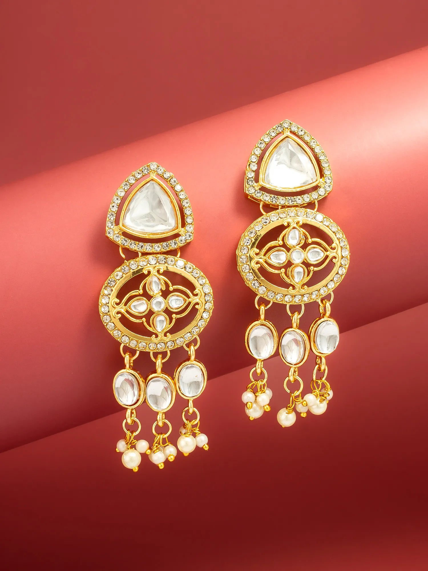 A pair of gold-toned traditional Indian dangle earrings with clear stones and pearl-like bead drops, displayed on a red background.