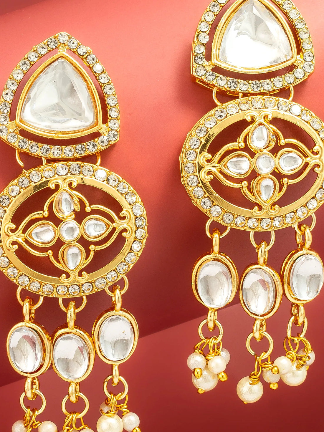 A pair of gold-toned traditional Indian dangle earrings with clear stones and pearl-like bead drops, displayed on a red background.