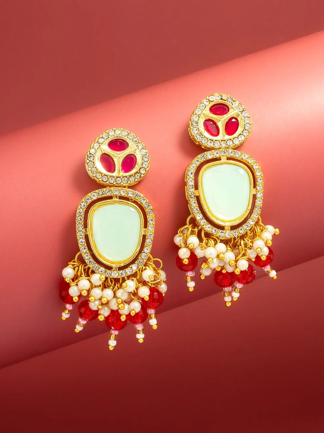 A pair of ornate gold-toned statement earrings with pale green stone centers, red gemstone accents, and dangling red and white bead chains, displayed on a curved pink-red background.