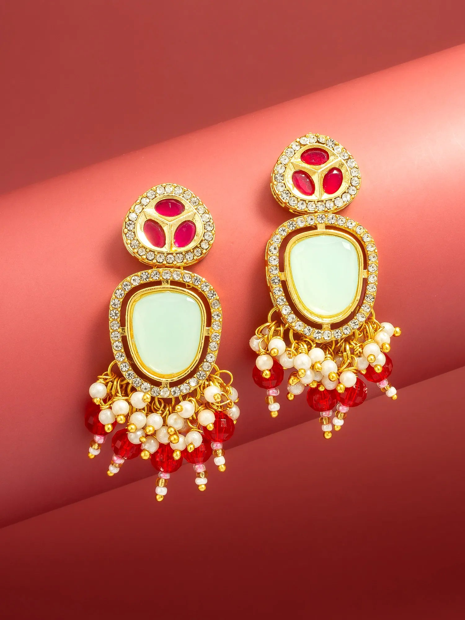 A pair of ornate gold-toned statement earrings with pale green stone centers, red gemstone accents, and dangling red and white bead chains, displayed on a curved pink-red background.