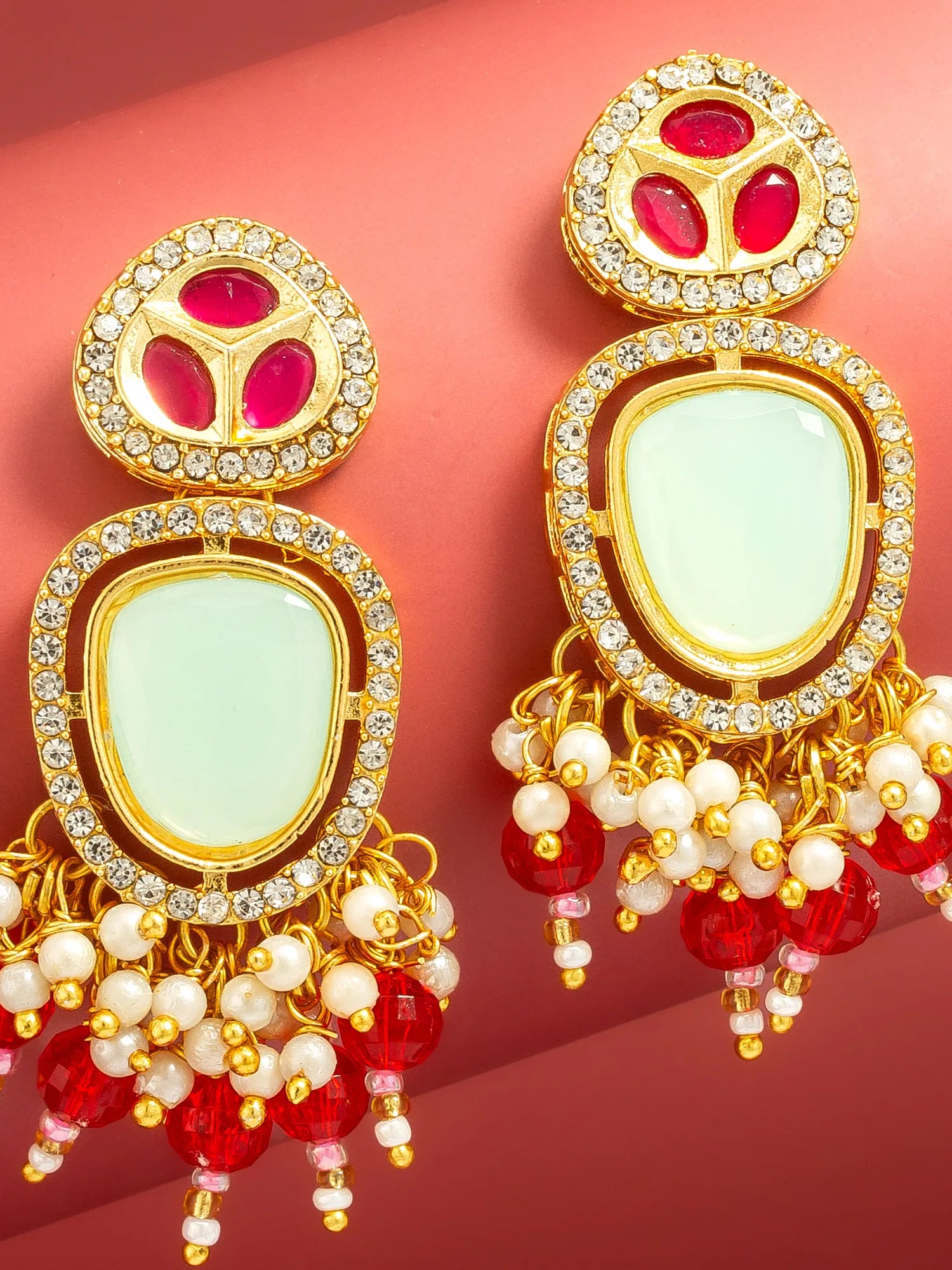 A pair of ornate gold-plated Indian jhumka earrings with light green stone centers, red stones at the top, and dangling red and white bead chains, set against a solid pink background.