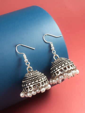A pair of silver-toned Indian jhumka earrings with dangling pearl beads, displayed on a blue cylindrical stand against a red background.