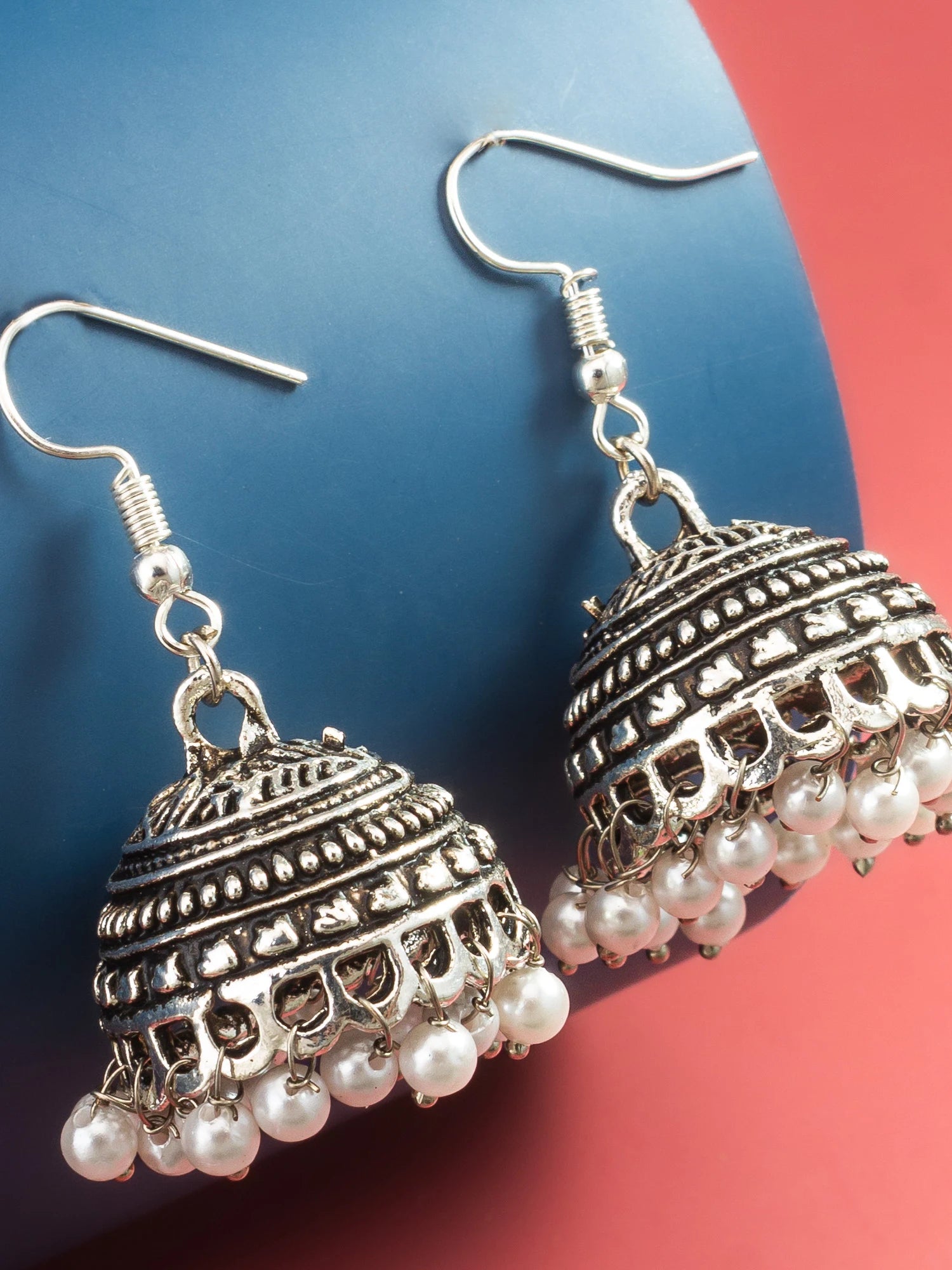 A pair of silver-toned Indian jhumka earrings with white pearl bead drops, displayed on a split blue and red background.