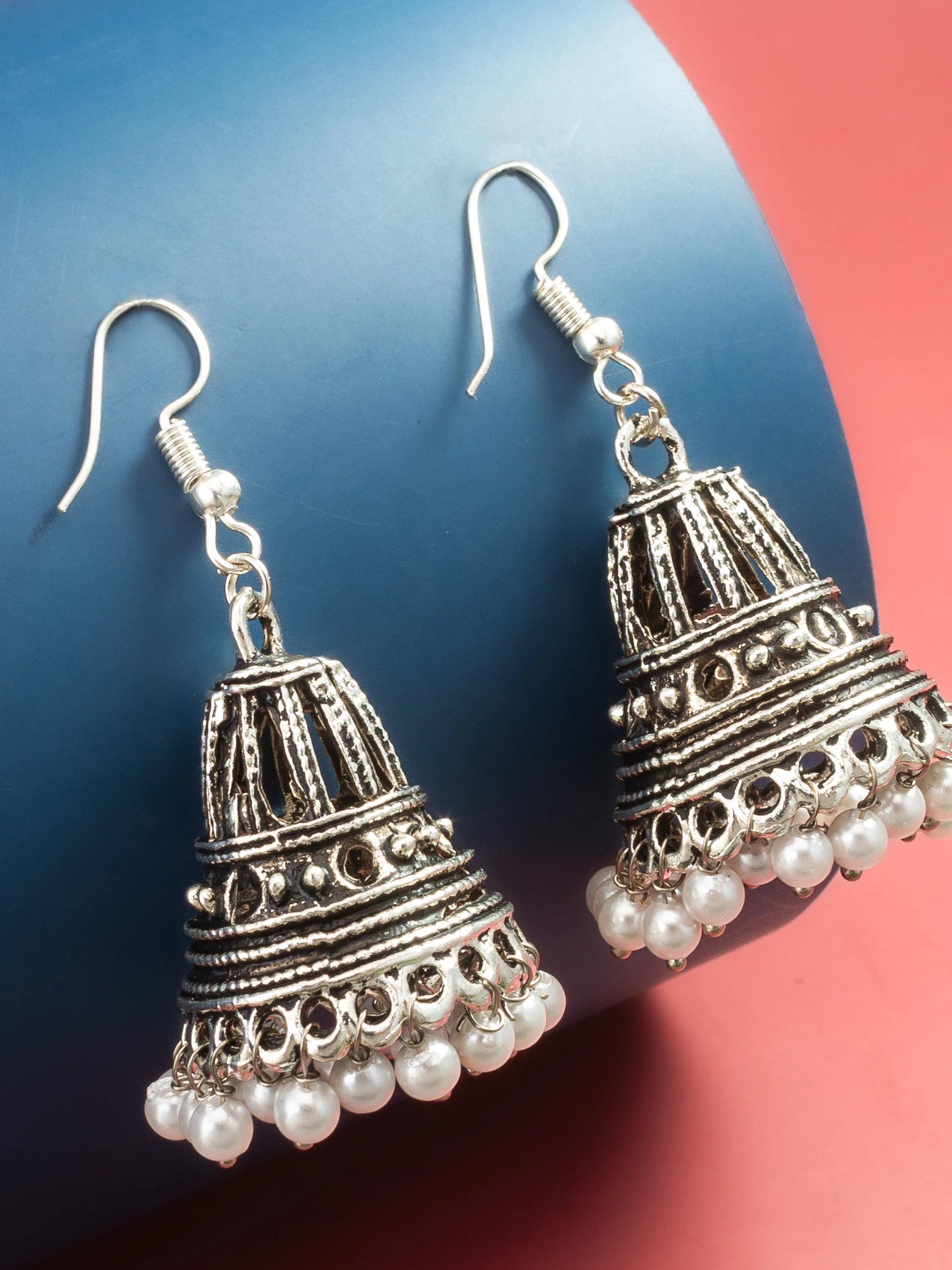 A pair of silver-toned bell-shaped jhumka earrings with dangling white pearl beads, displayed on a curved blue and red background.