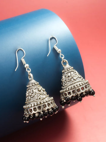 A pair of silver-toned filigree jhumka earrings with small black bead drops, displayed on a blue cylindrical stand against a pink background.