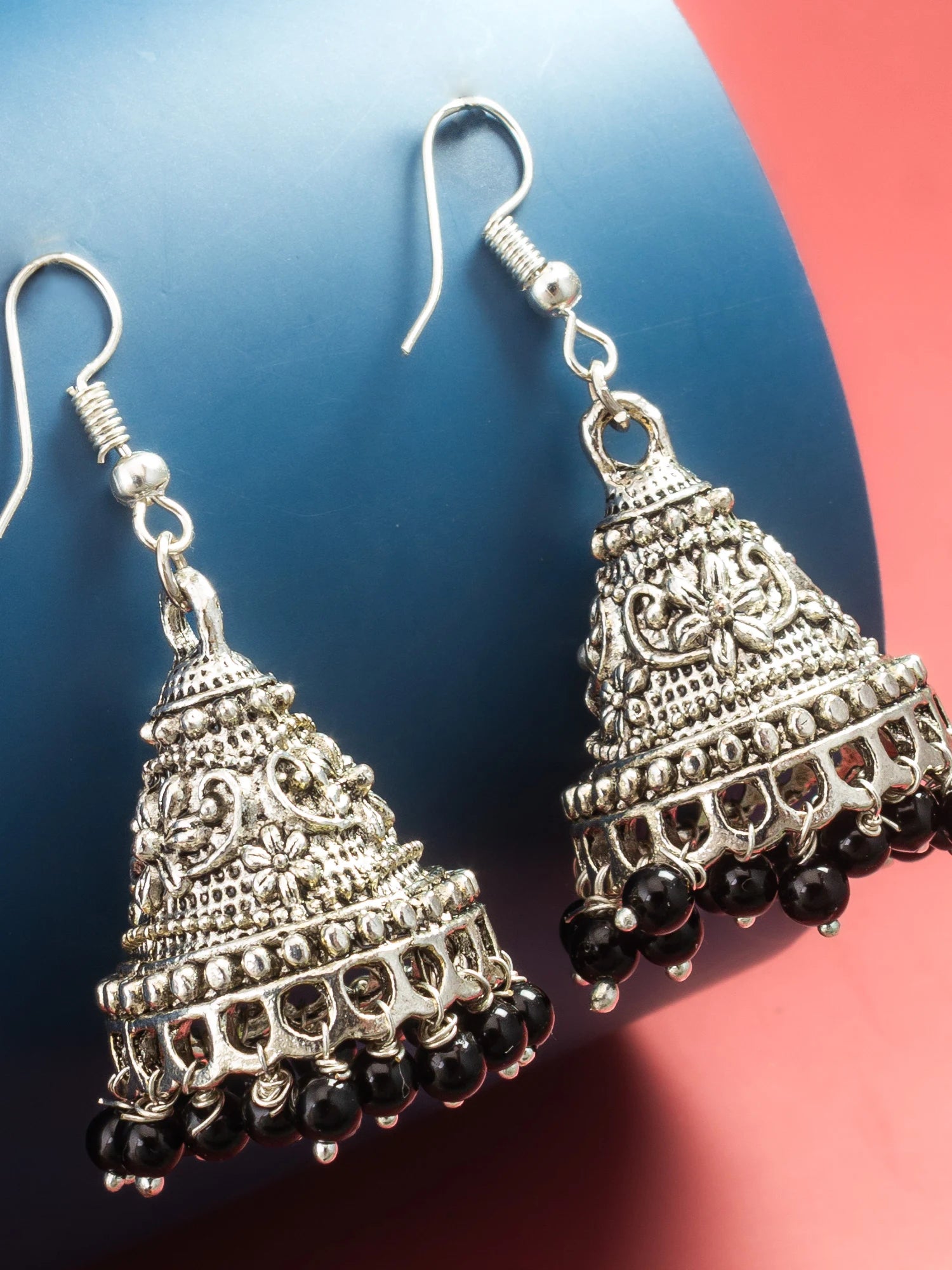 A pair of silver-toned Indian jhumka earrings with intricate filigree work and dangling black beads, displayed on a split blue and red background.