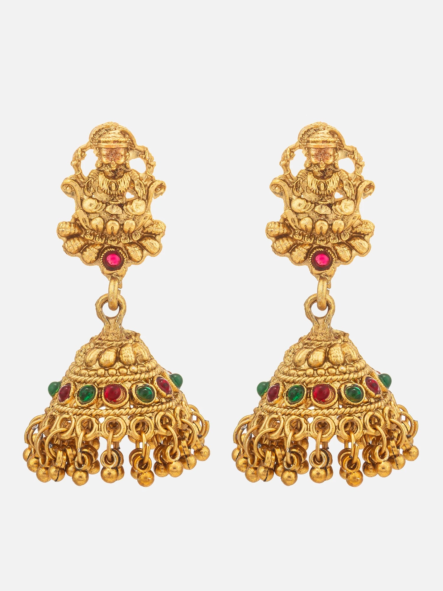 A pair of ornate gold-toned jhumka earrings with detailed deity motifs at the top and dangling bell-shaped charms, accented with small red and green stones, shown on a light gray background.