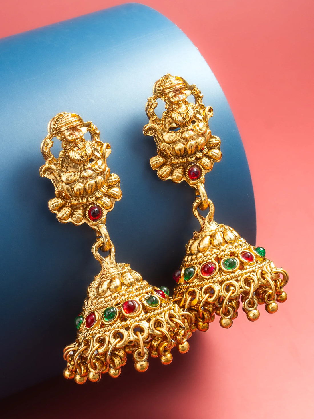 A pair of ornate gold-toned jhumka earrings with red and green stone accents, displayed on a blue cylindrical prop against a pink background.