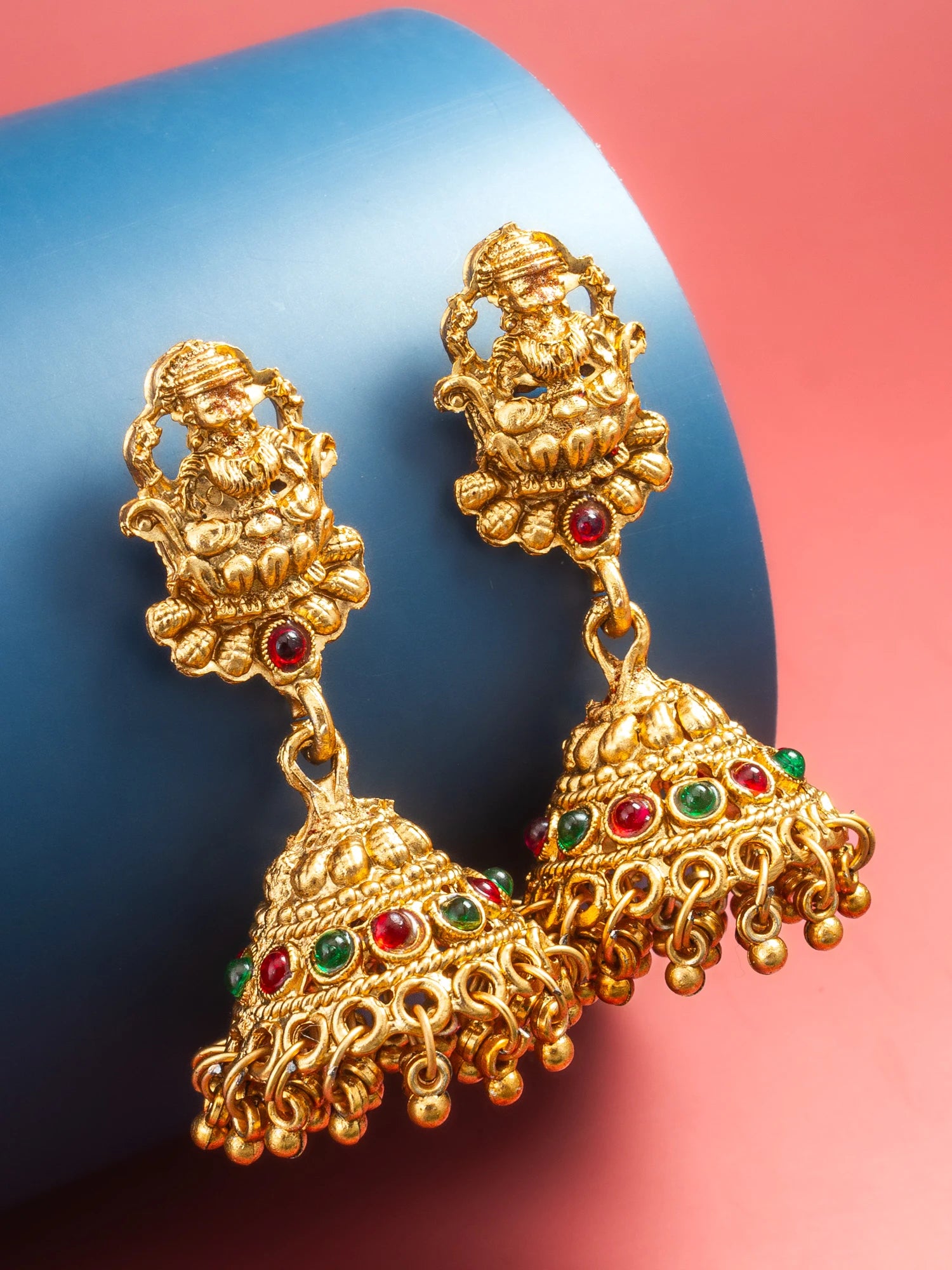 A pair of ornate gold-toned jhumka earrings with red and green stone accents, displayed on a blue cylindrical prop against a pink background.