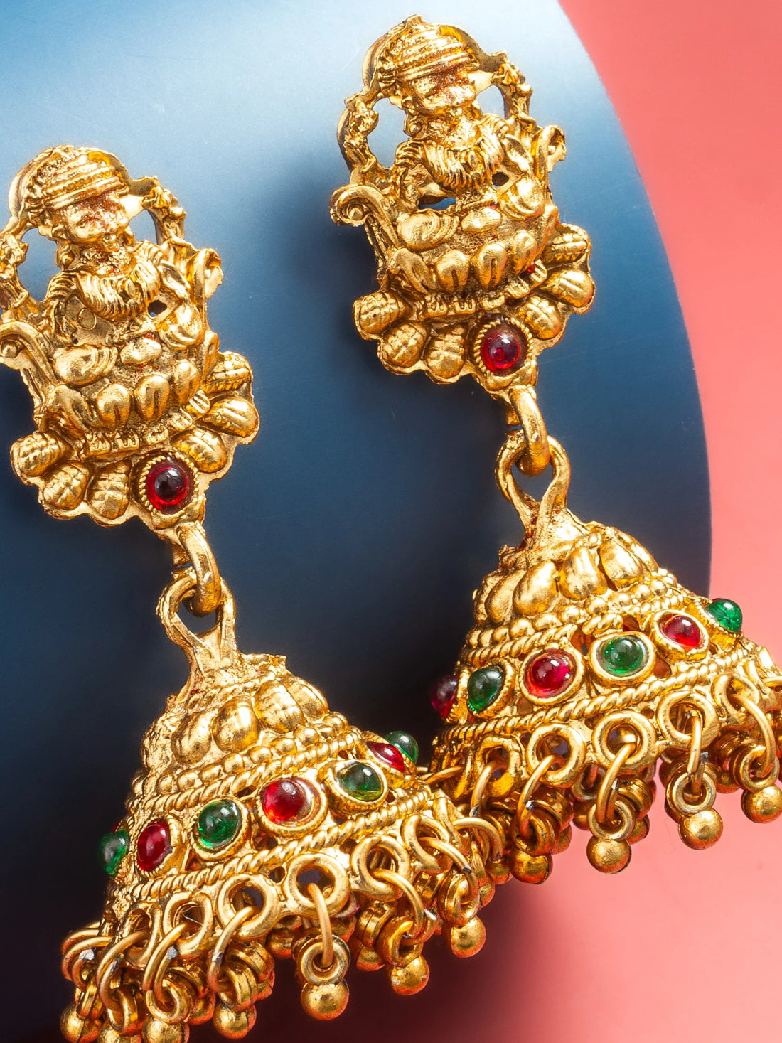 A pair of ornate gold-toned jhumka earrings with red and green stone accents, displayed on a blue cylindrical prop against a pink background.