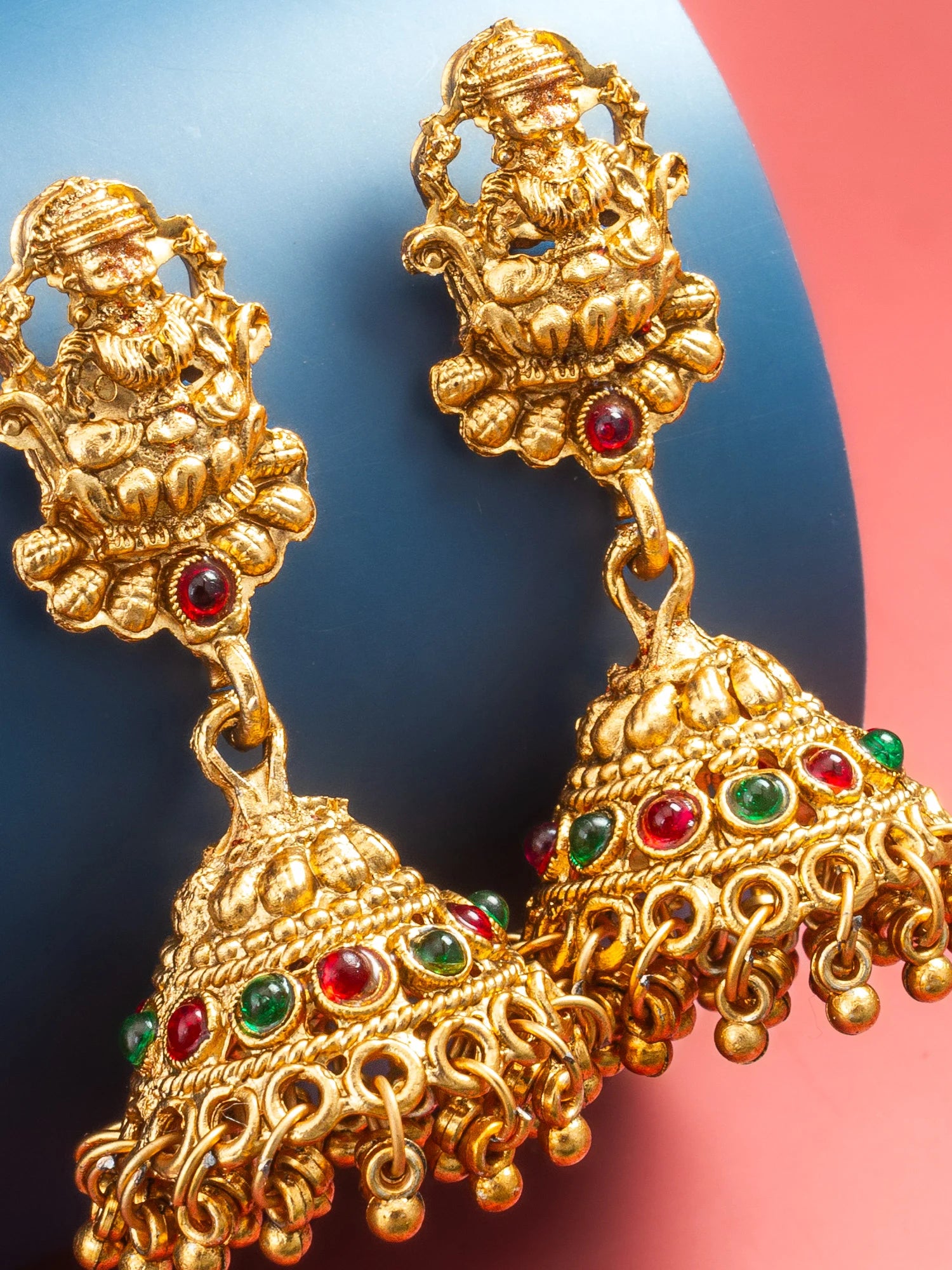 A pair of ornate gold-toned Indian jhumka earrings with detailed deity motifs and small red and green gemstone accents, shown against a split blue and pink background.