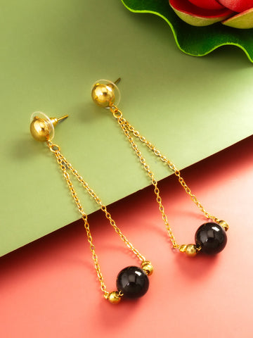 Gold-tone dangle earrings with spherical black beads and small gold spacer beads, displayed on a split green and coral background.