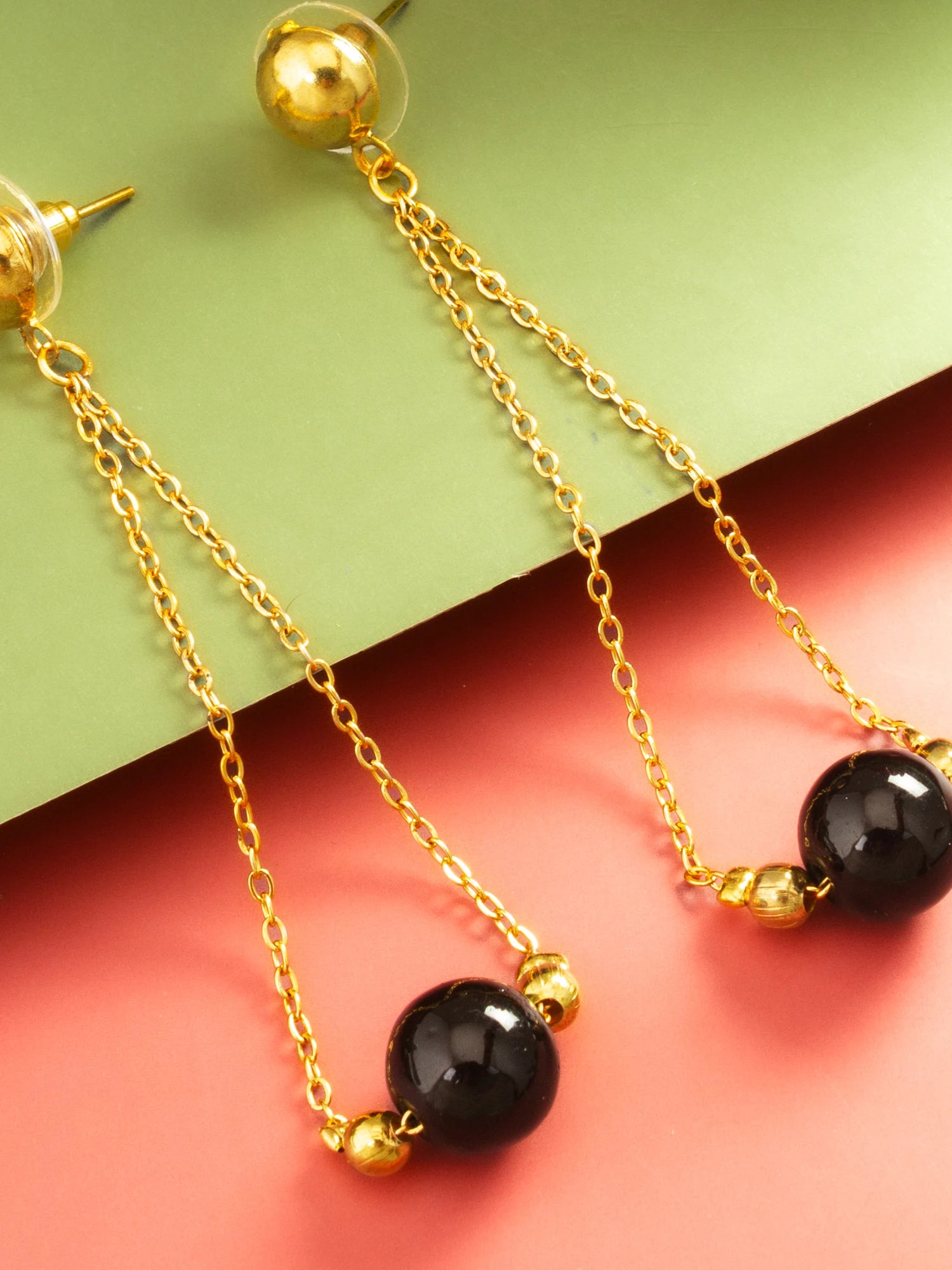 Gold dangle earrings with black round beads and small gold spacer beads, displayed on a split green and coral background.