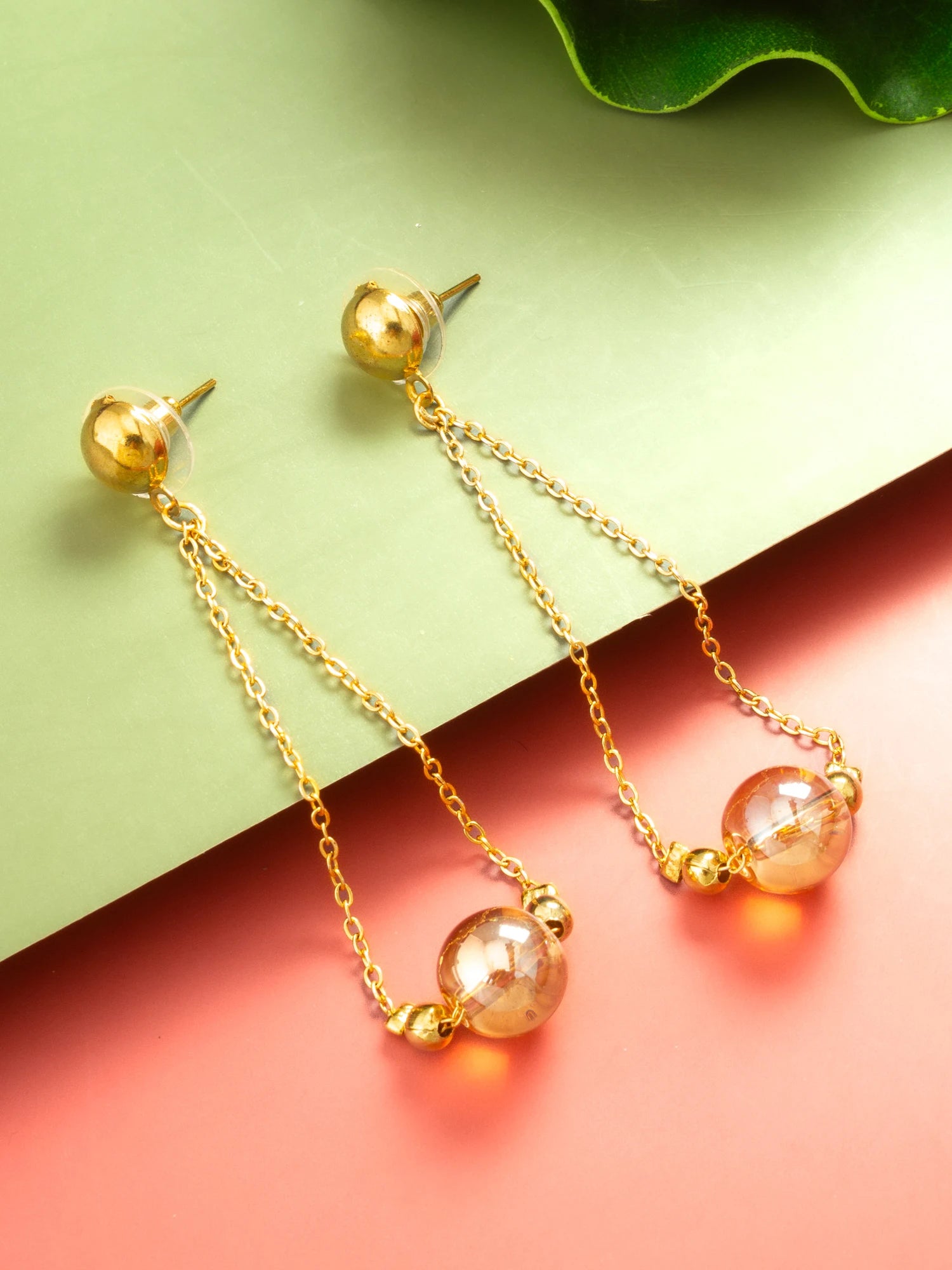 A pair of gold dangle earrings with spherical gold studs, thin chain drops, and clear faceted beads that catch the light, displayed on a split green and coral background.