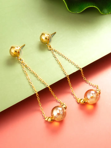 A pair of gold dangle earrings with spherical gold studs, thin chain drops, and clear faceted beads that catch the light, displayed on a split green and coral background.