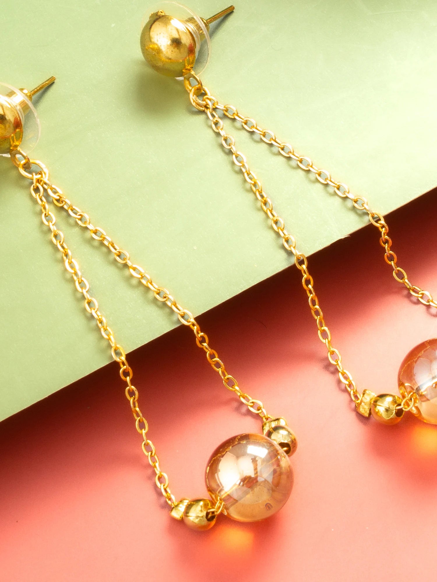 Gold dangle earrings with clear faceted glass beads and small gold bell charms, displayed on a split green and pink background.