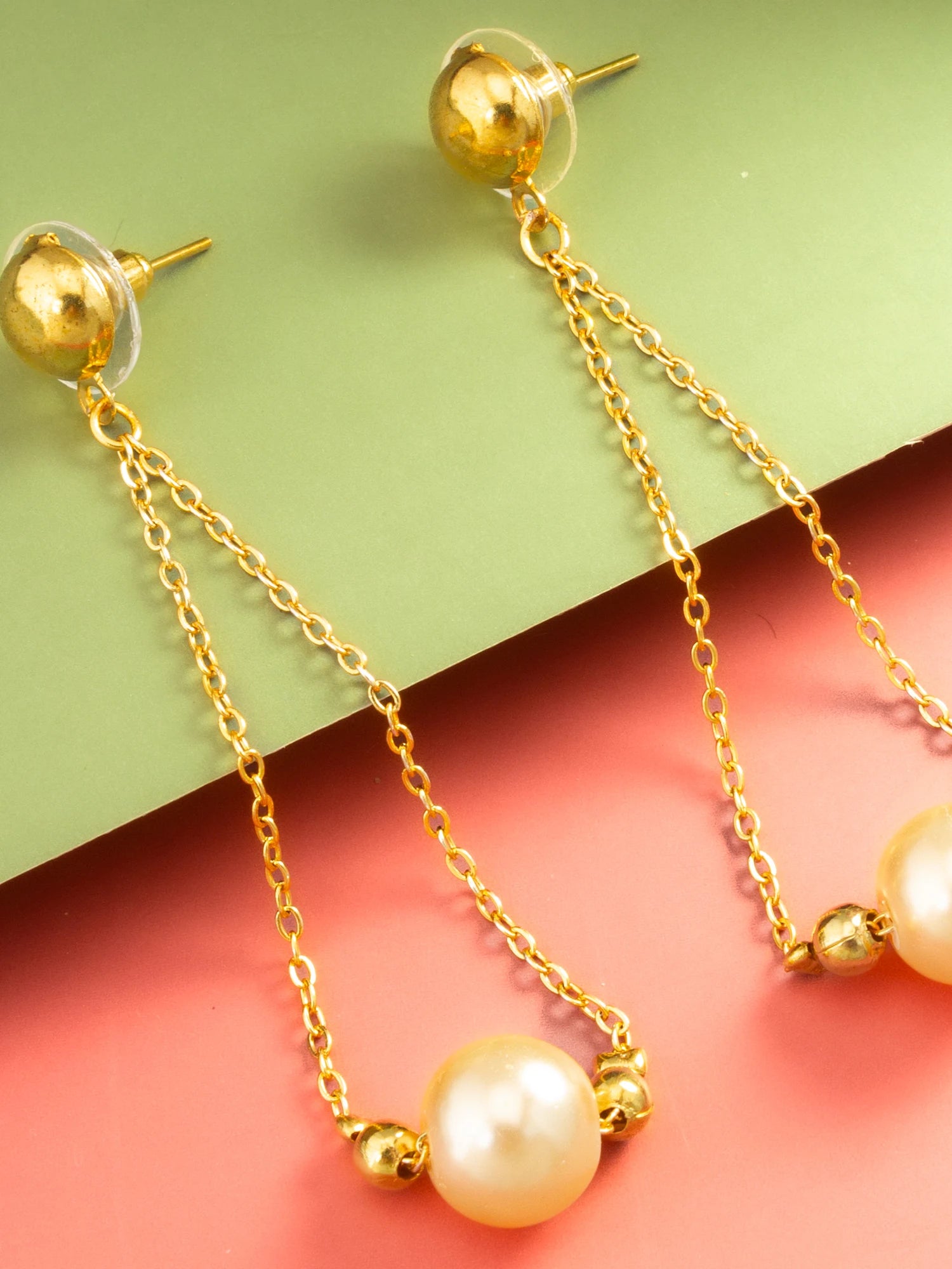Gold dangle earrings with spherical gold posts, short chain links, small gold beads, and large cream pearl drops, displayed on a split green and coral background.