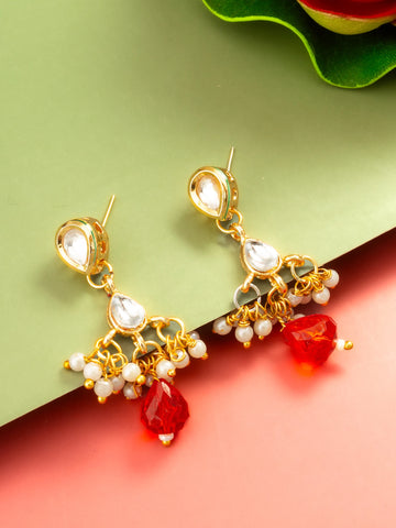 Gold-plated jhumka earrings with white stones and red teardrop beads, displayed on a split green and pink background.