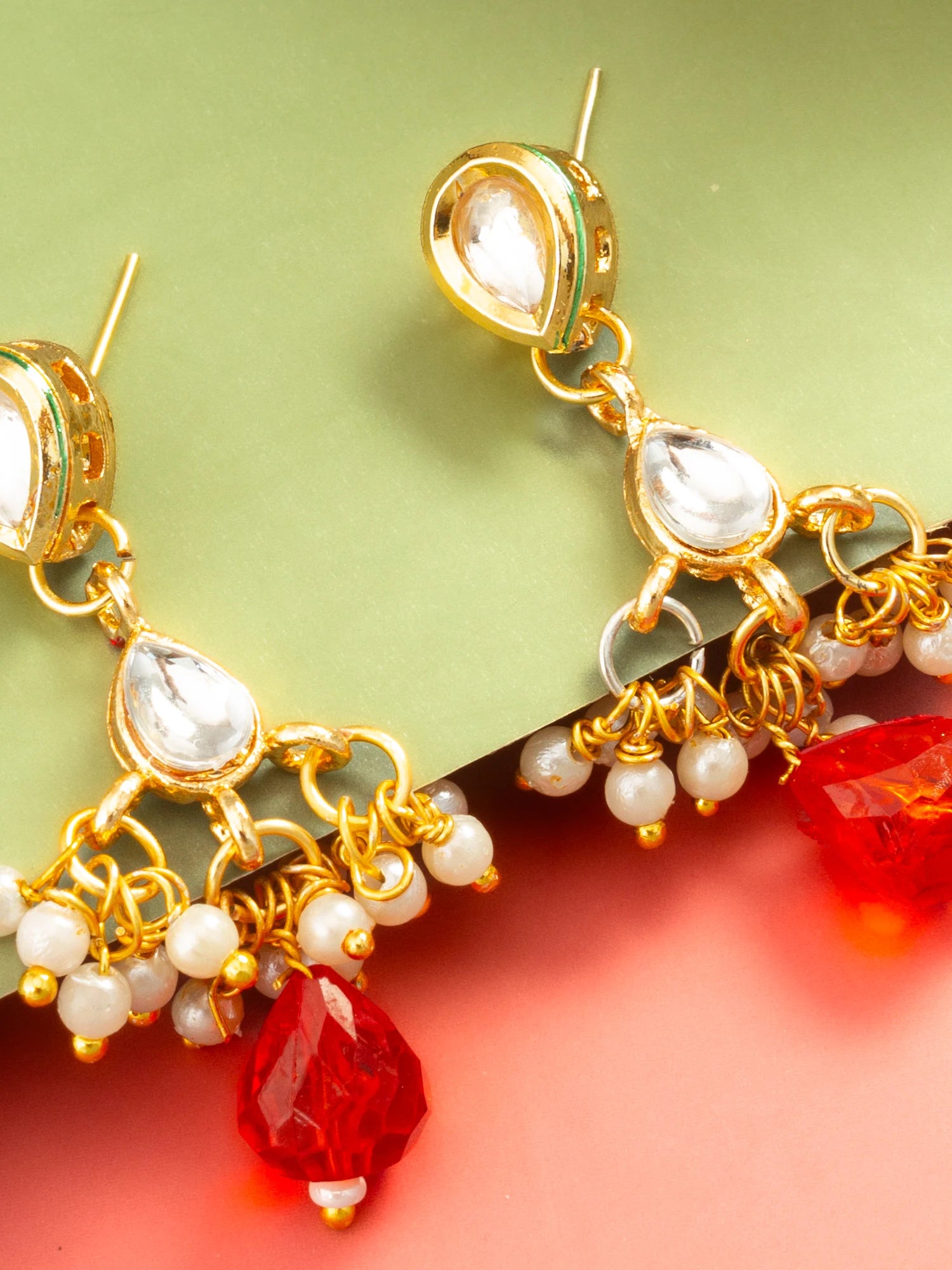 Gold-plated Indian jhumka earrings with white stone accents, white pearl beads, and red faceted crystal drops, shown on a split green and pink background.