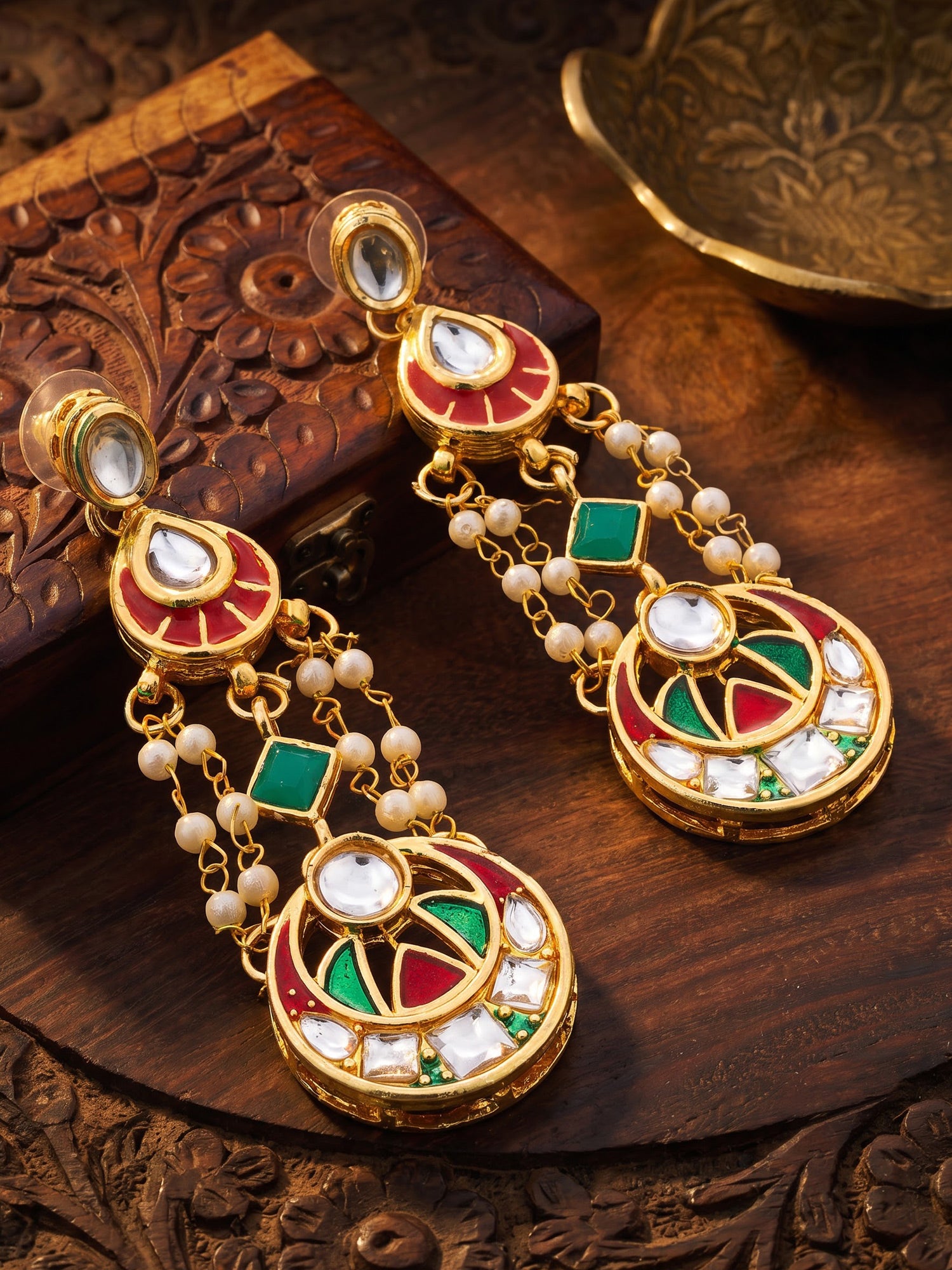 A pair of ornate gold-plated Indian dangle earrings decorated with red, green, and white enamel, white stones, and small white pearls, displayed on a carved wooden surface.