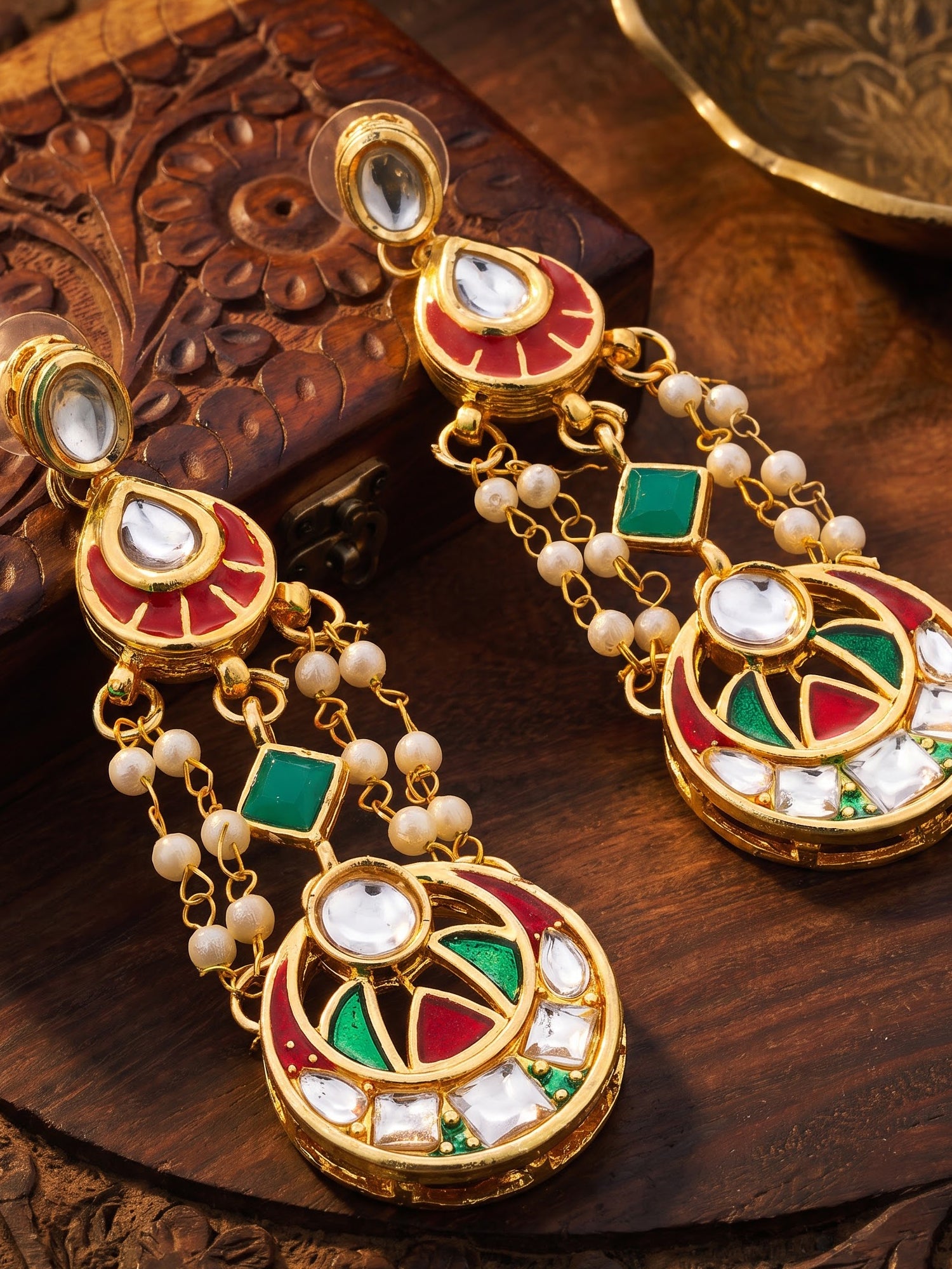 A pair of ornate gold-plated Indian dangle earrings decorated with red, green, and white enamel, white stones, and small white pearls, displayed on a carved wooden surface.