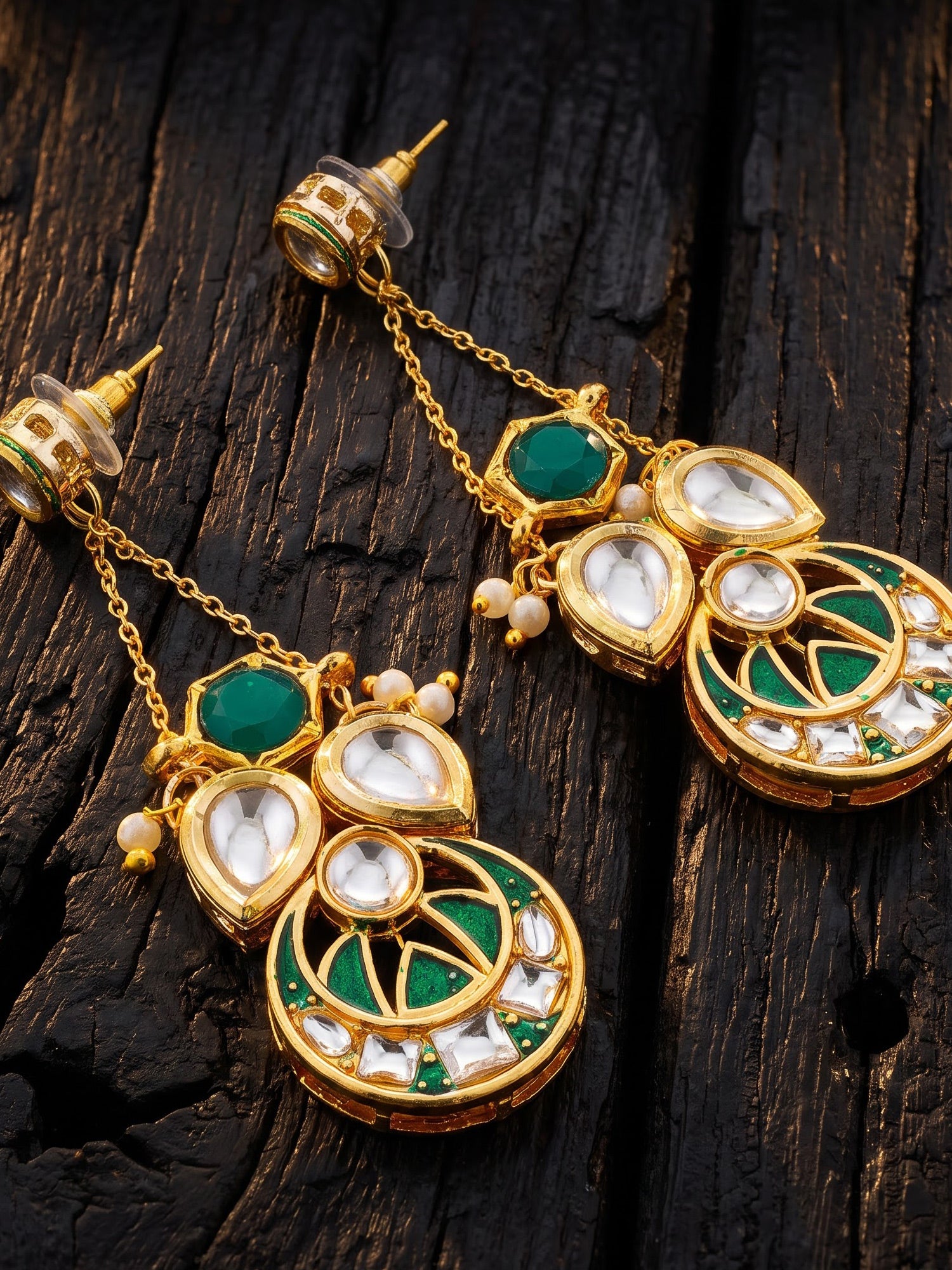 Gold-plated Indian Kundan dangle earrings with green enamel, white stones, and small pearl accents, displayed on a dark textured wooden surface.