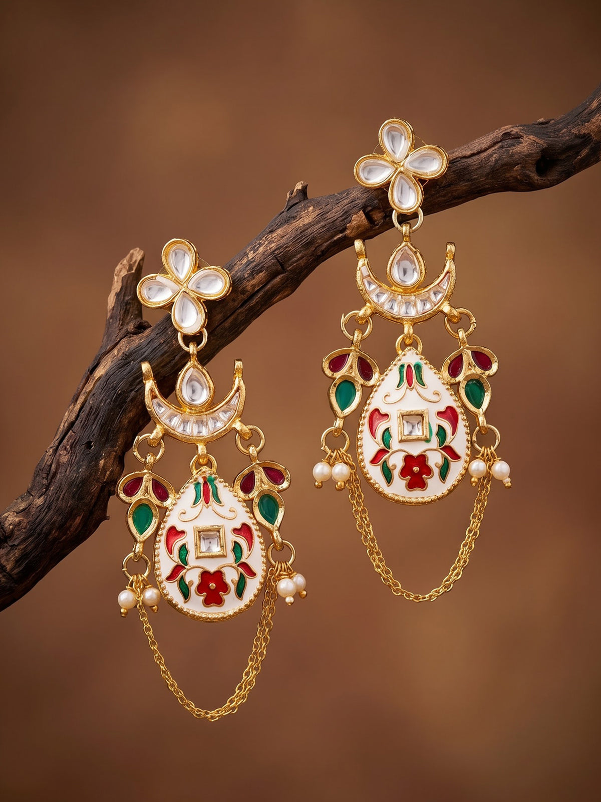 A pair of ornate gold-toned Indian jhumka earrings with white stone floral tops, crescent moon details, and white enamel pendants decorated with red and green floral enameled motifs, hanging from a dark brown branch against a warm brown background.